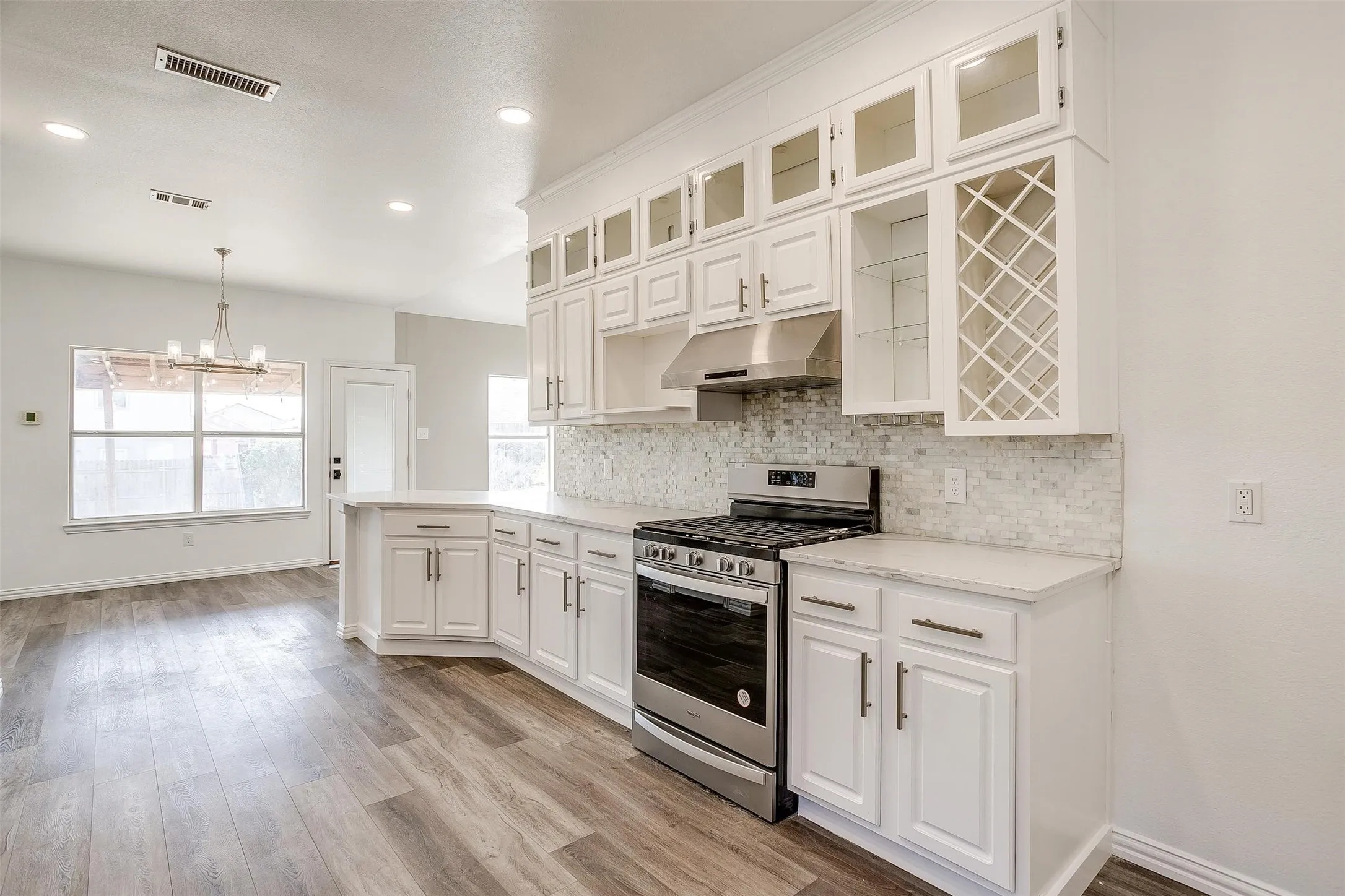 Bright, open kitchen featuring white cabinetry with glass-front upper cabinets, stainless steel gas range, quartz countertops, marble subway tile backsplash, and wood-finish flooring throughout the space