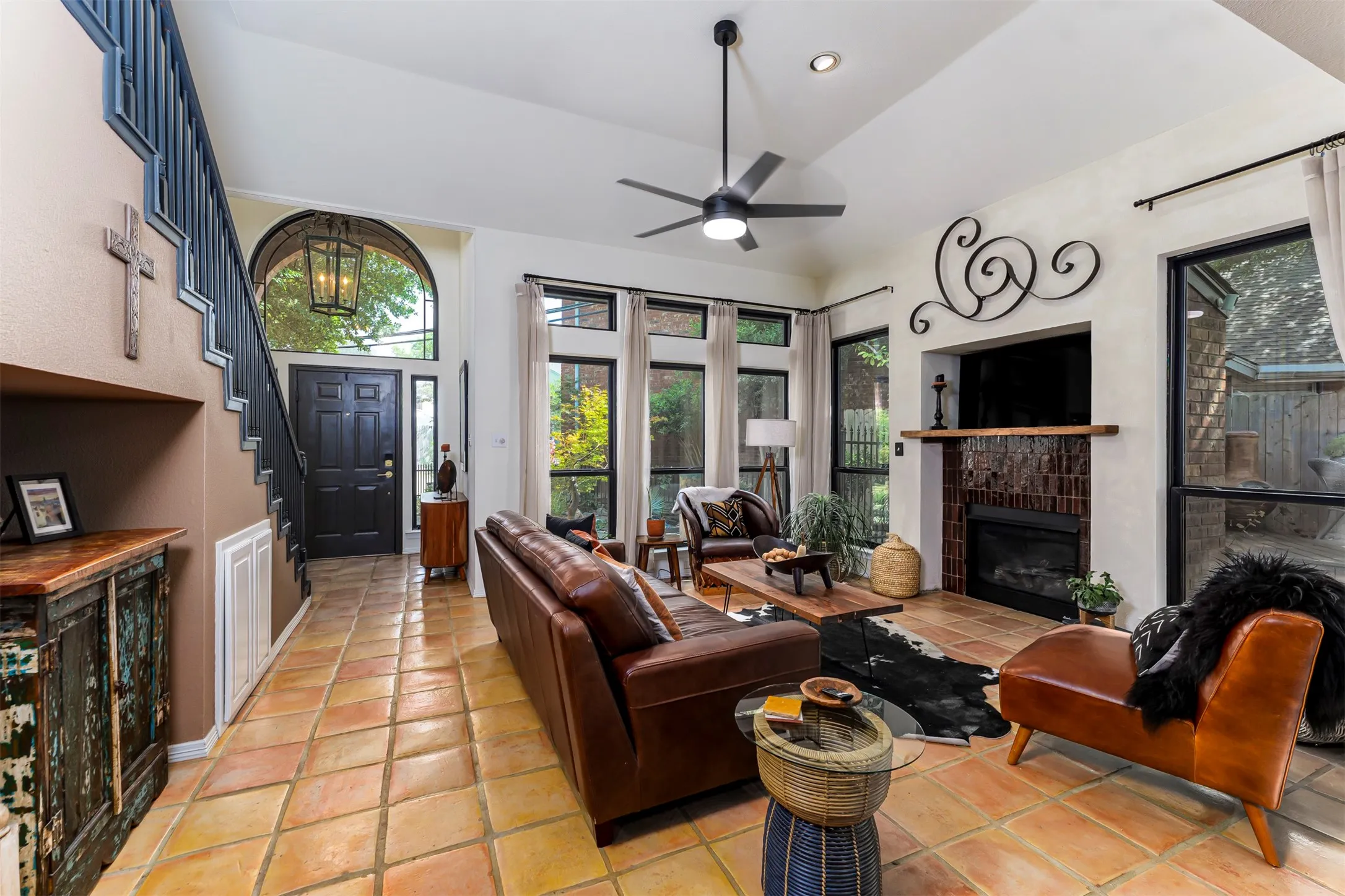 Vaulted ceiling with a ceiling fan, tiled flooring, built-in fireplace with a wooden mantel, multiple large windows, and a black staircase with an arched window above the entry door
