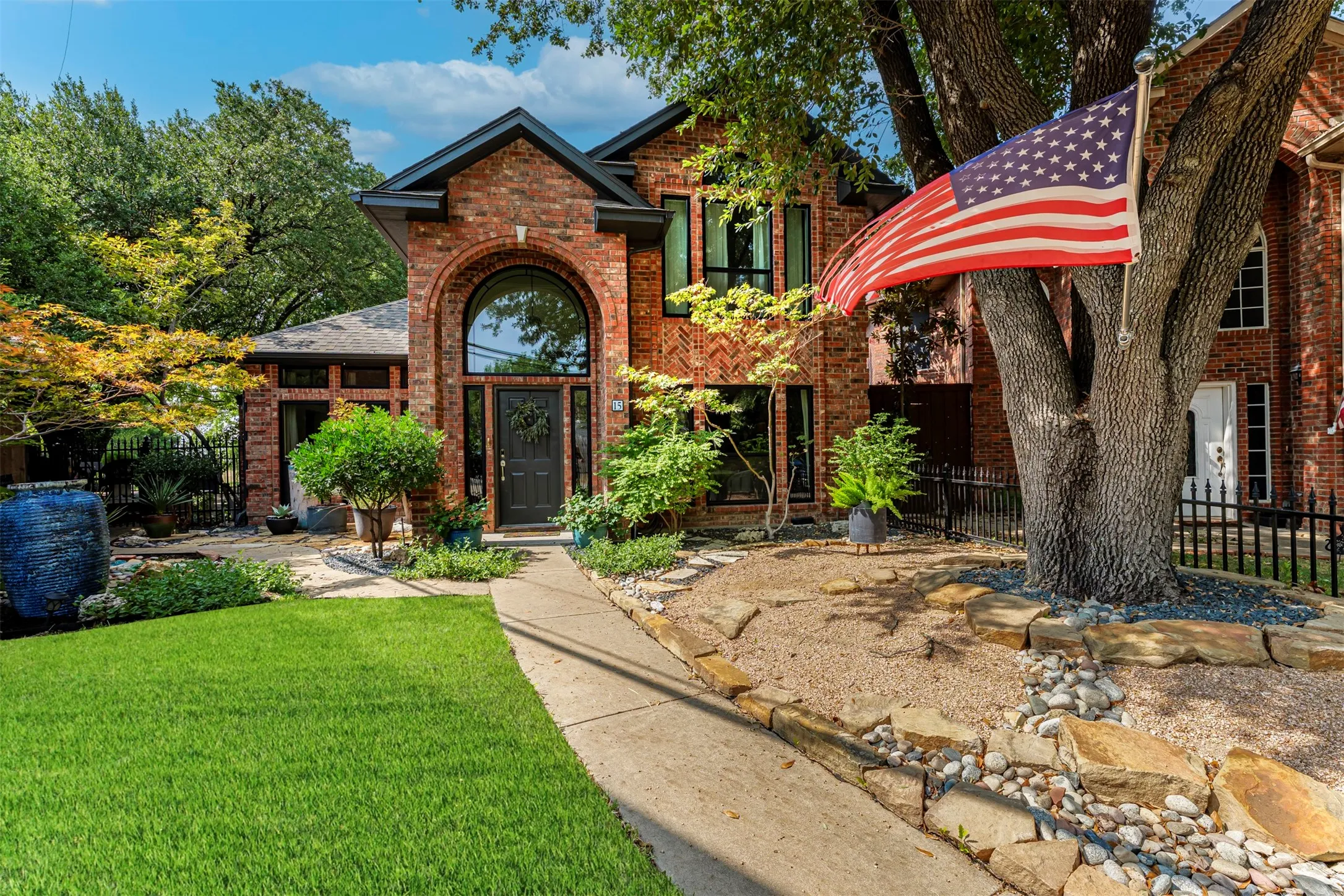 Brick exterior with a prominent arched entry, large picture windows, mature trees, a landscaped front yard, and a paved walkway