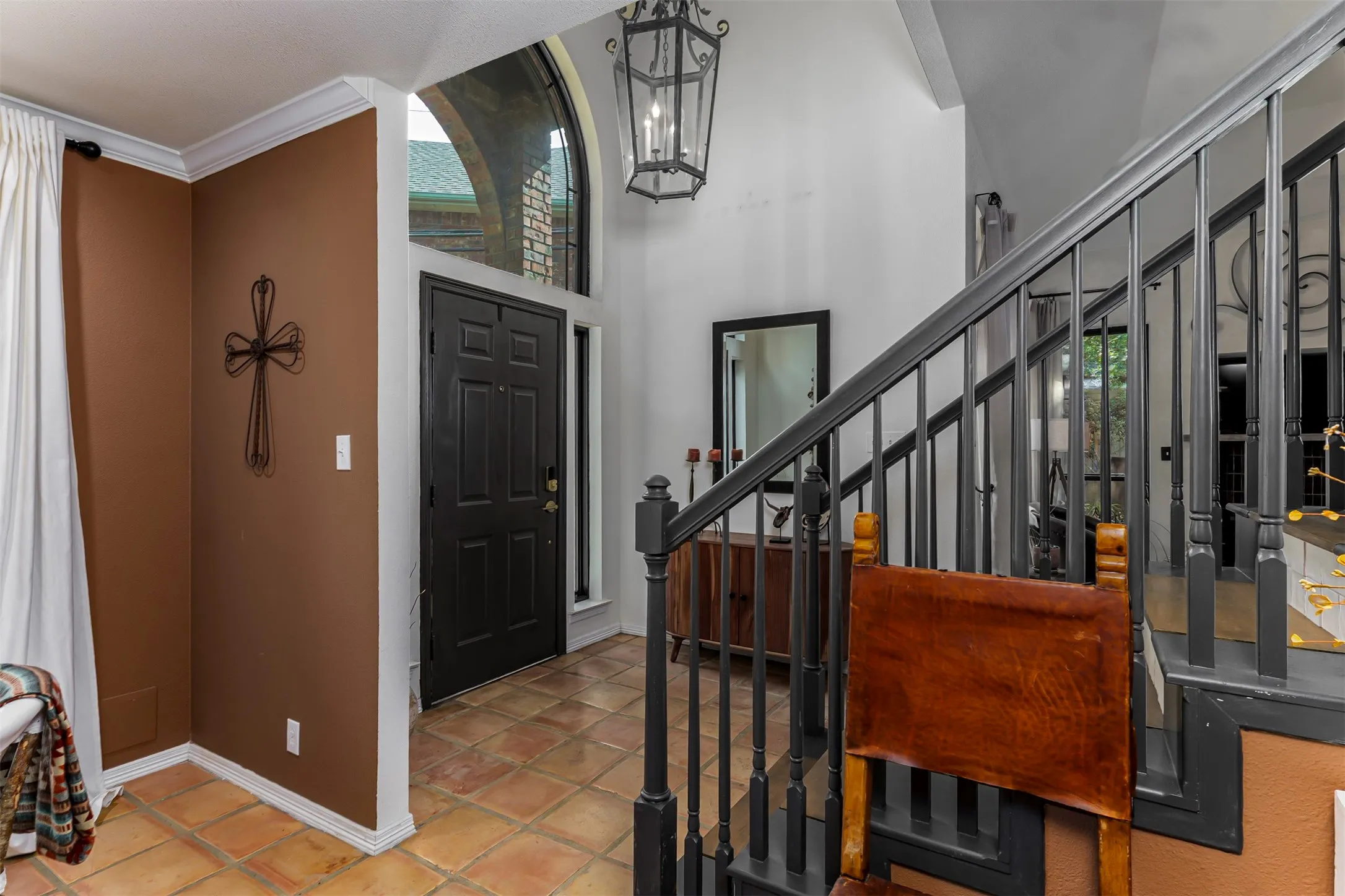 Welcoming foyer showcasing terracotta tile flooring, warm earth-tone walls, ornamental cross accent, and a striking staircase with wood-finish railings and metal balusters