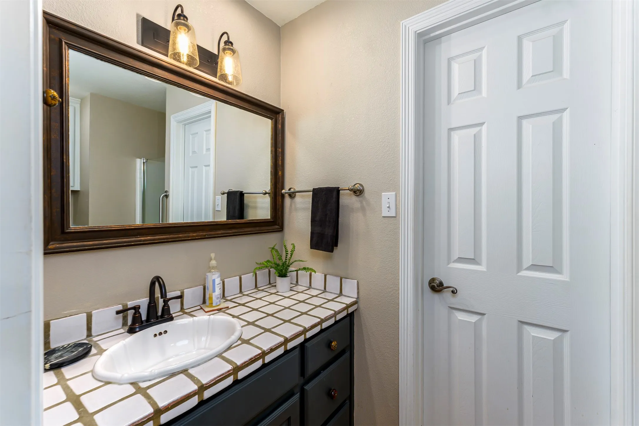 Elegant primary bathroom featuring a large framed mirror with bronze hardware, dual vanity with black cabinetry and geometric tile backsplash, polished black faucet, and brass sconce lighting above