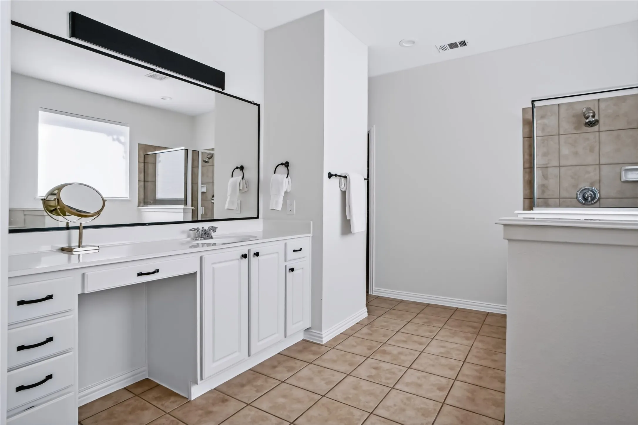 White vanity with integrated sink, expansive mirror, tile flooring, and a tiled shower enclosure
