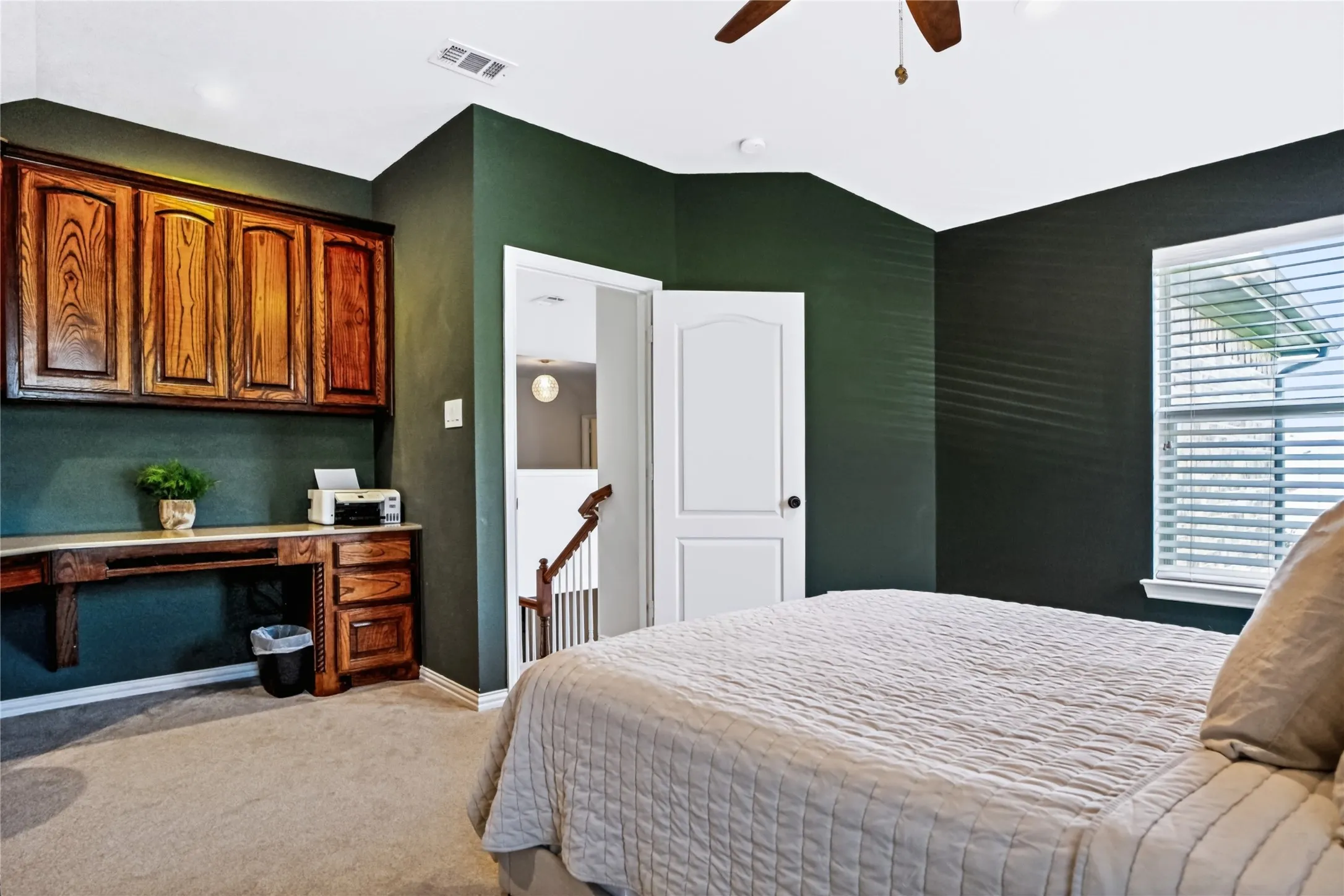 Spacious primary bedroom featuring deep forest green accent walls, hardwood cabinetry with upper storage, a dedicated work desk area, carpet flooring, plantation shutters, and a ceiling fan