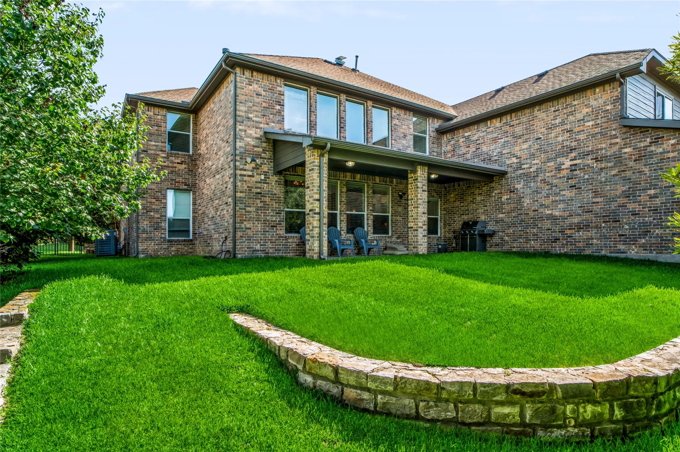 Expansive backyard featuring a manicured lawn, stone retaining wall, and a covered patio with brick columns
