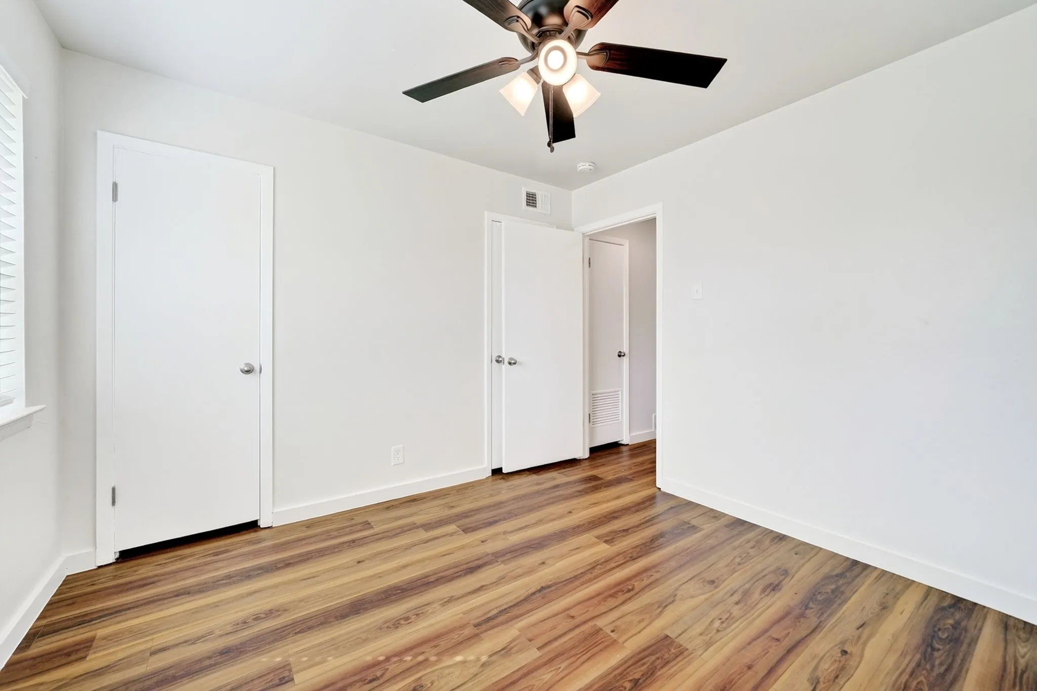 Wood-finish flooring, white walls, ceiling fan, white door, and window with blinds