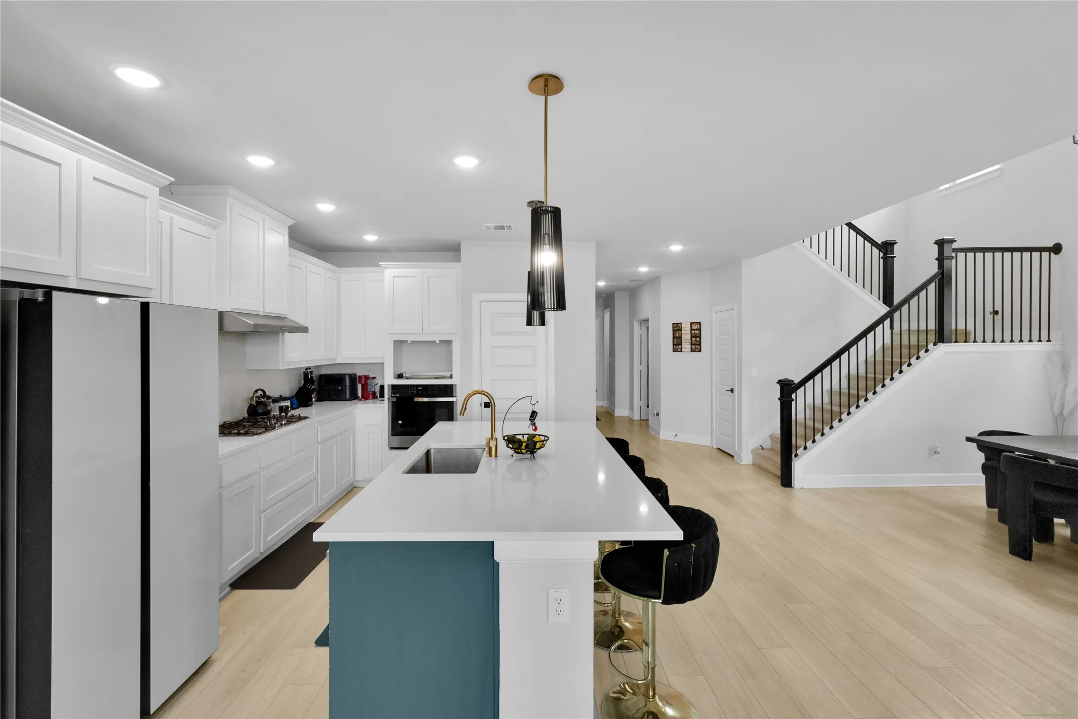 Open-concept kitchen and living area featuring white cabinetry, stainless steel appliances, a large white quartz island with teal base, recessed and pendant lighting, wood-finish flooring, and a grand staircase with black metal railings