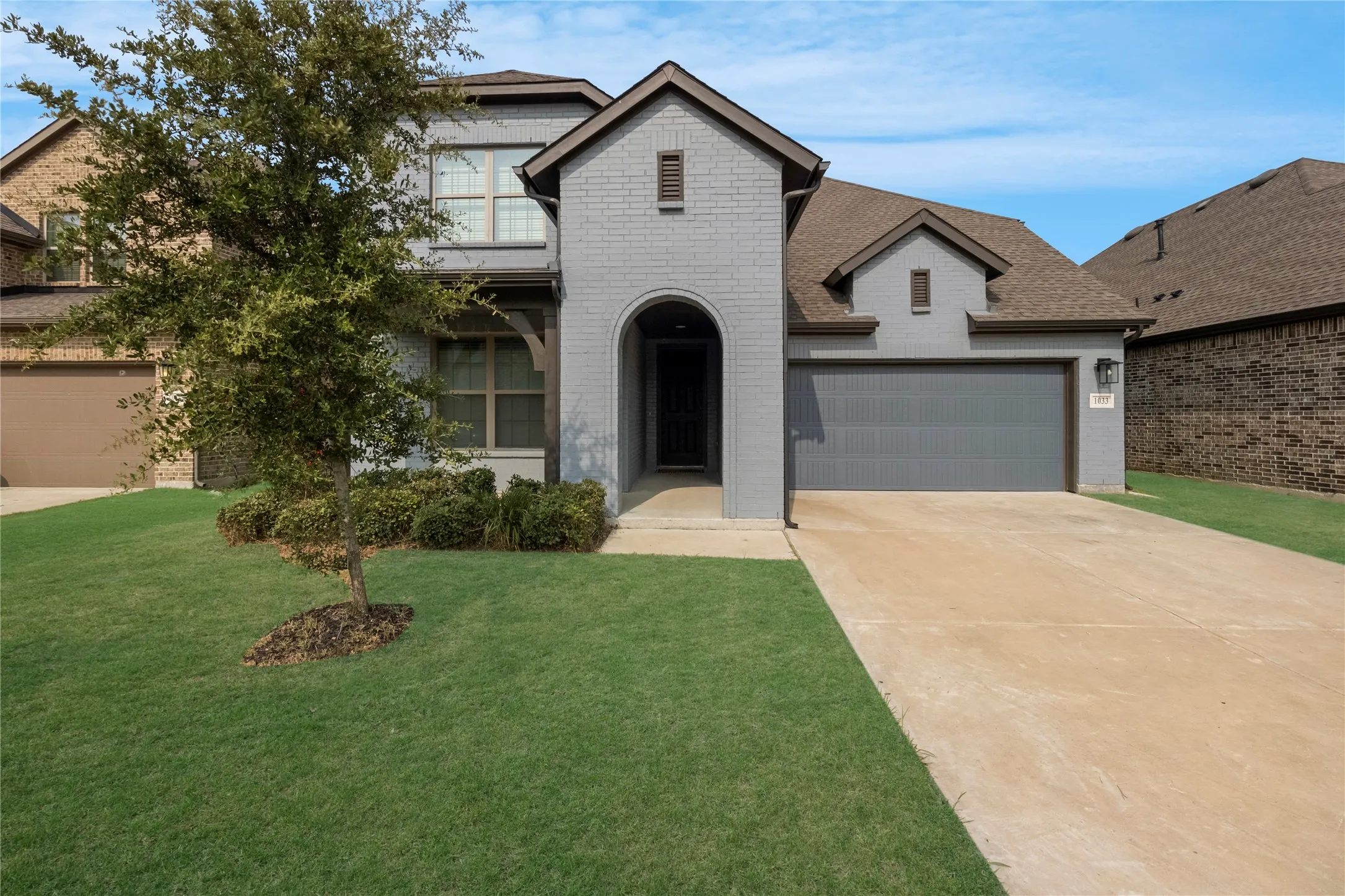 Contemporary two-story residence featuring a gray painted brick exterior, an arched entry, a two-car garage, and a lush front lawn with mature landscaping