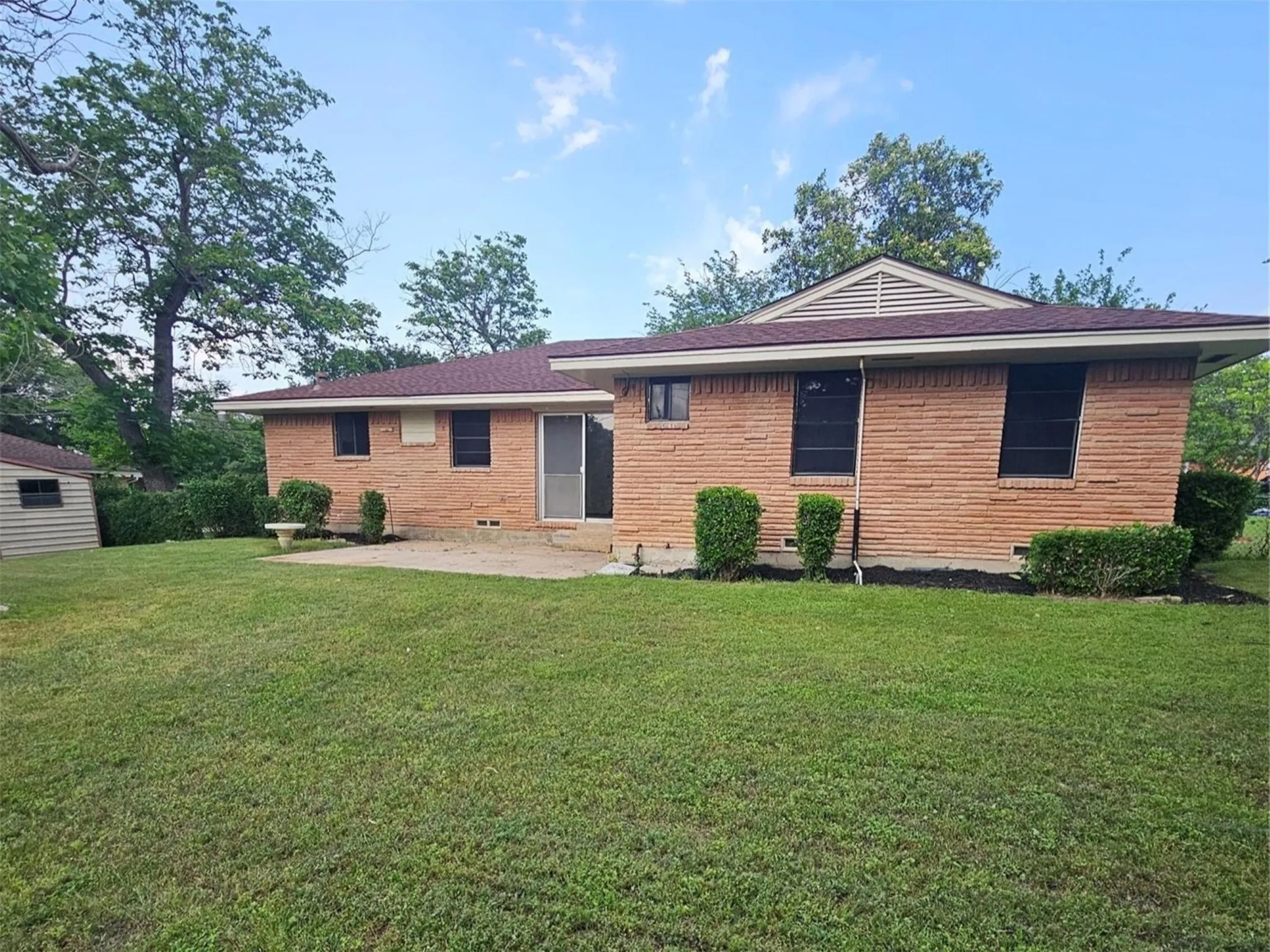 Expansive backyard featuring a brick exterior, concrete patio, mature trees, and a storage shed
