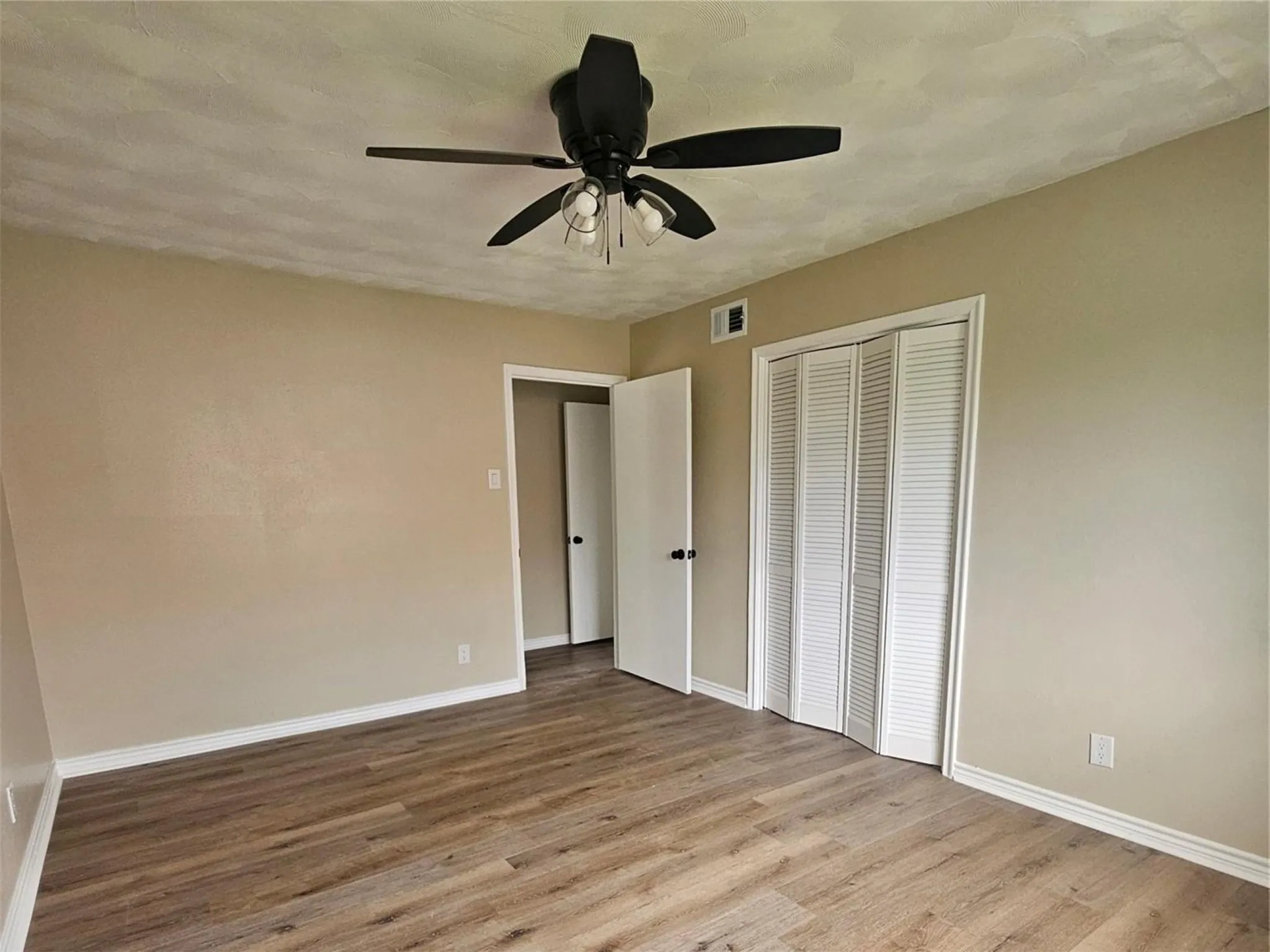Spacious bedroom featuring wood-finish flooring, black matte ceiling fan with integrated lighting, and generous closet space with louvered bi-fold doors