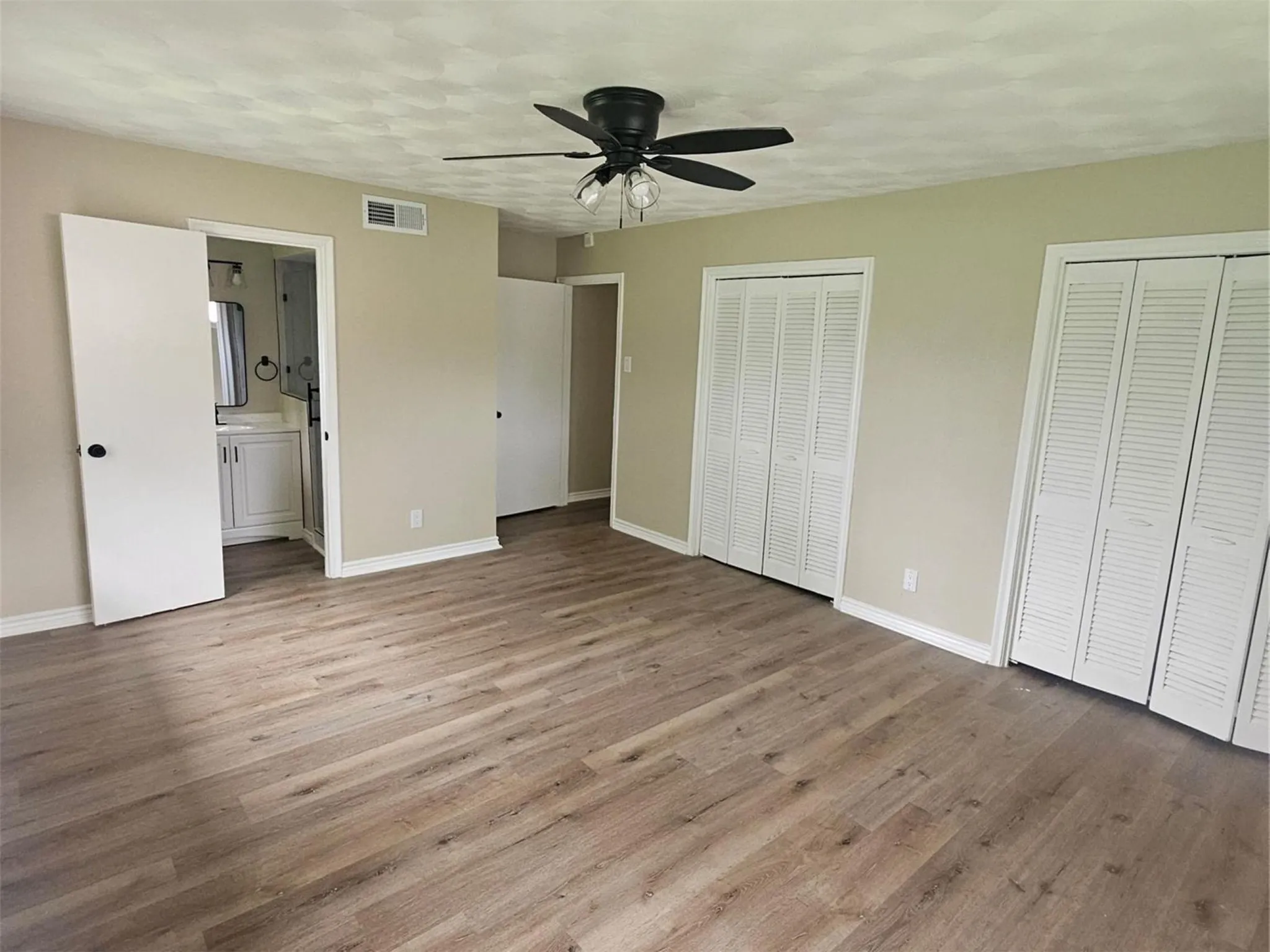 Spacious primary bedroom featuring 2 closets, private bathroom, wood-finish flooring, natural light, neutral wall paint, black matte finished ceiling fan with integrated lighting