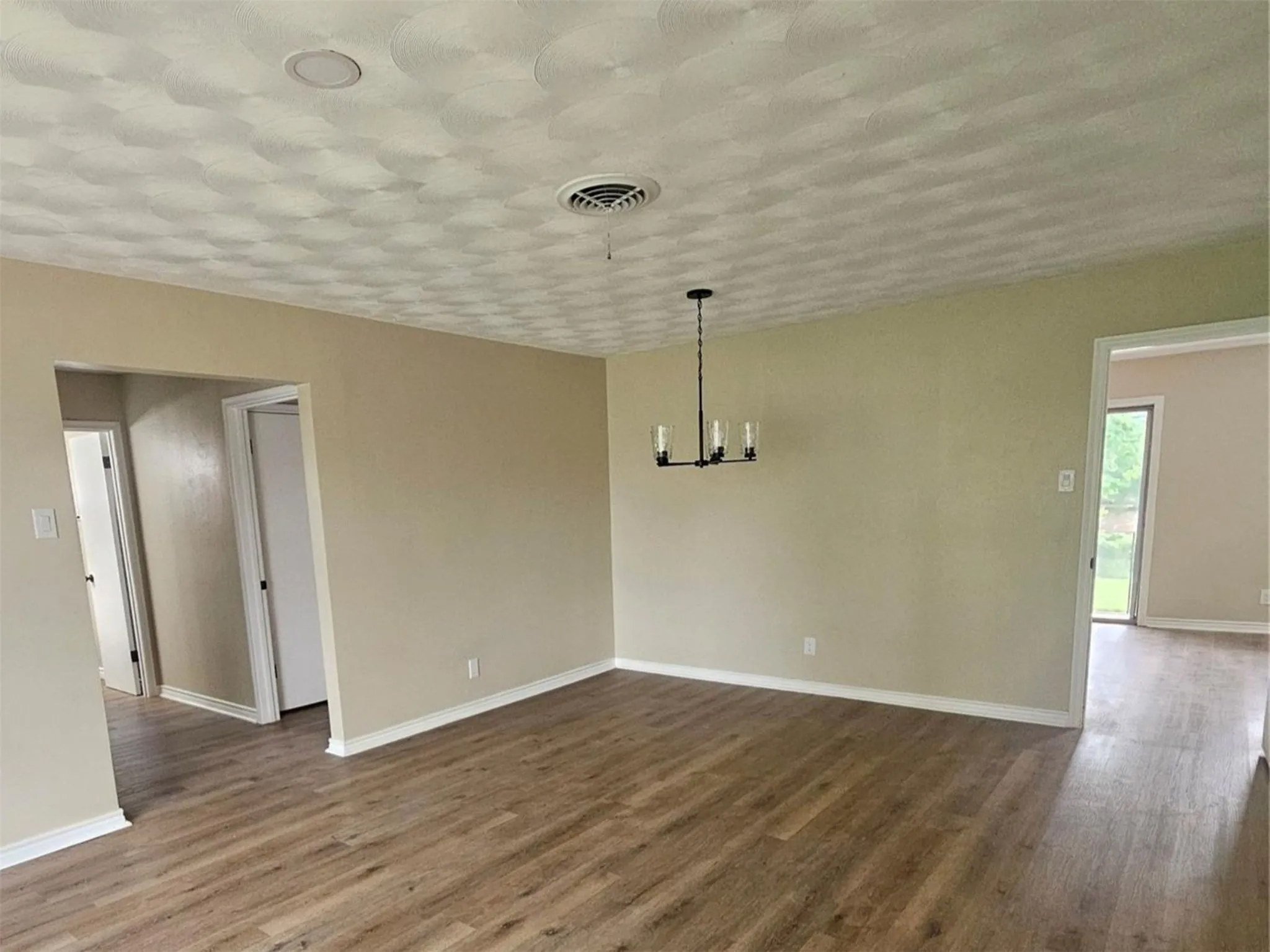 Spacious room featuring wood-finish flooring, natural light, neutral wall paint, black matte finished chandelier and crisp white baseboards