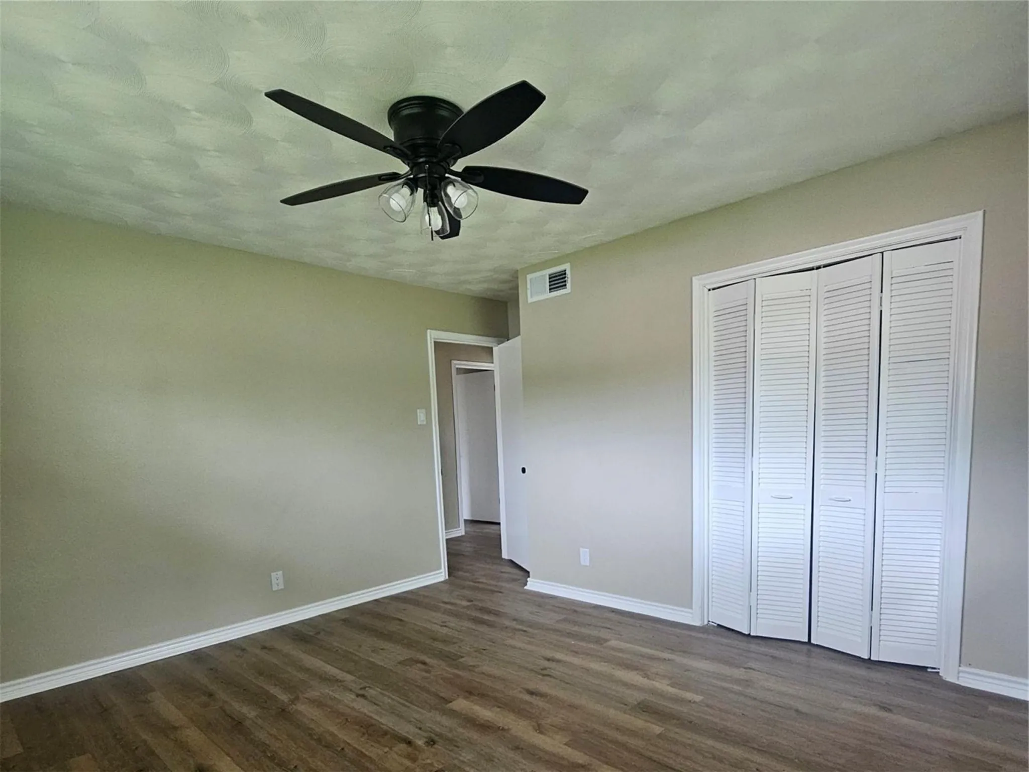 Spacious bedroom featuring wood-finish flooring, black matte ceiling fan with integrated lighting, and generous closet space with louvered bi-fold doors