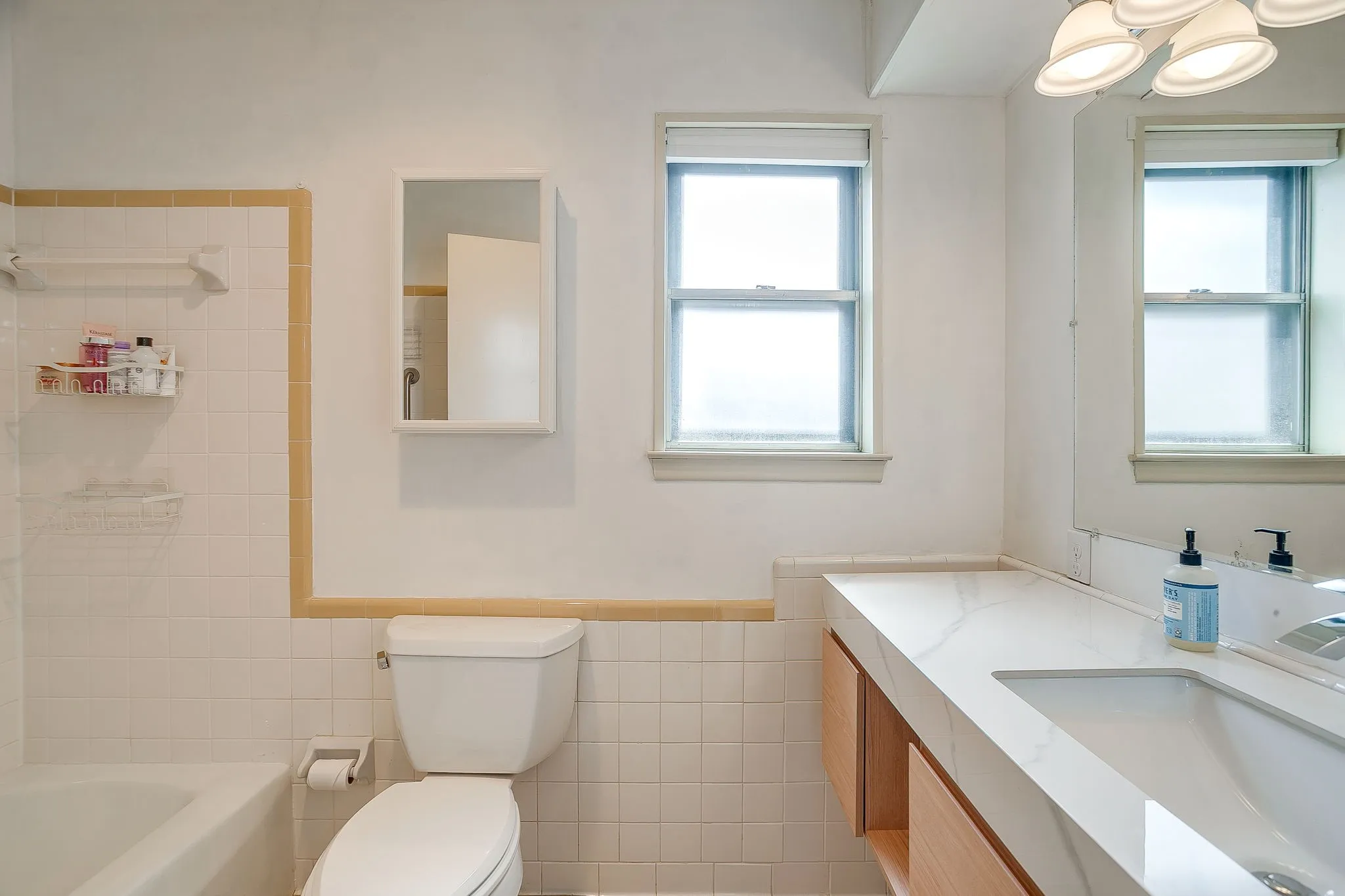 White tiled walls with decorative trim, built-in medicine cabinet, double-hung windows, integrated vanity sink, and light wood-finish cabinetry