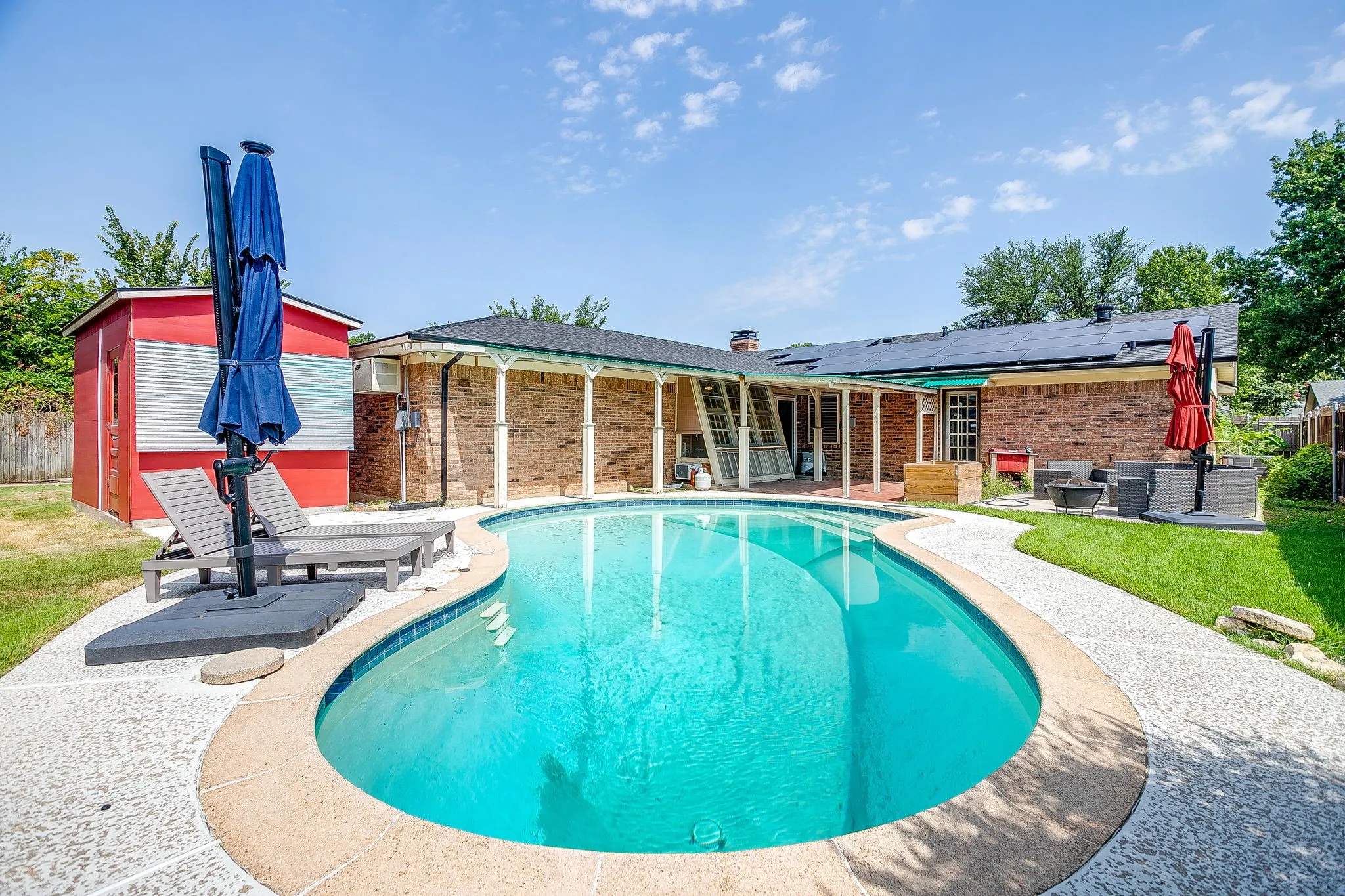 Freeform swimming pool with a concrete surround, brick exterior, covered patio, and a red storage shed