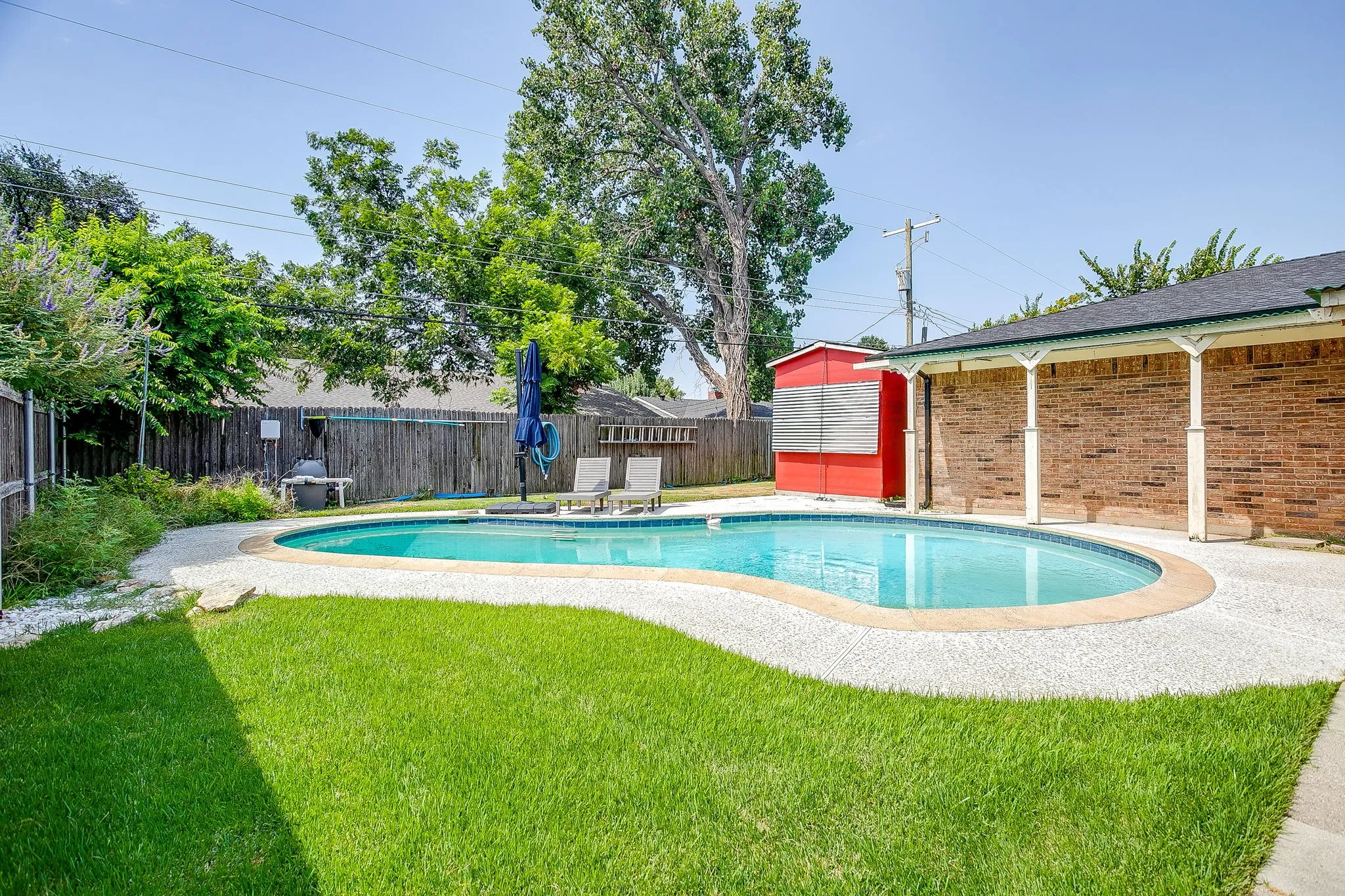 Swimming pool with surrounding concrete decking, a brick exterior wall with a covered patio, and a red shed