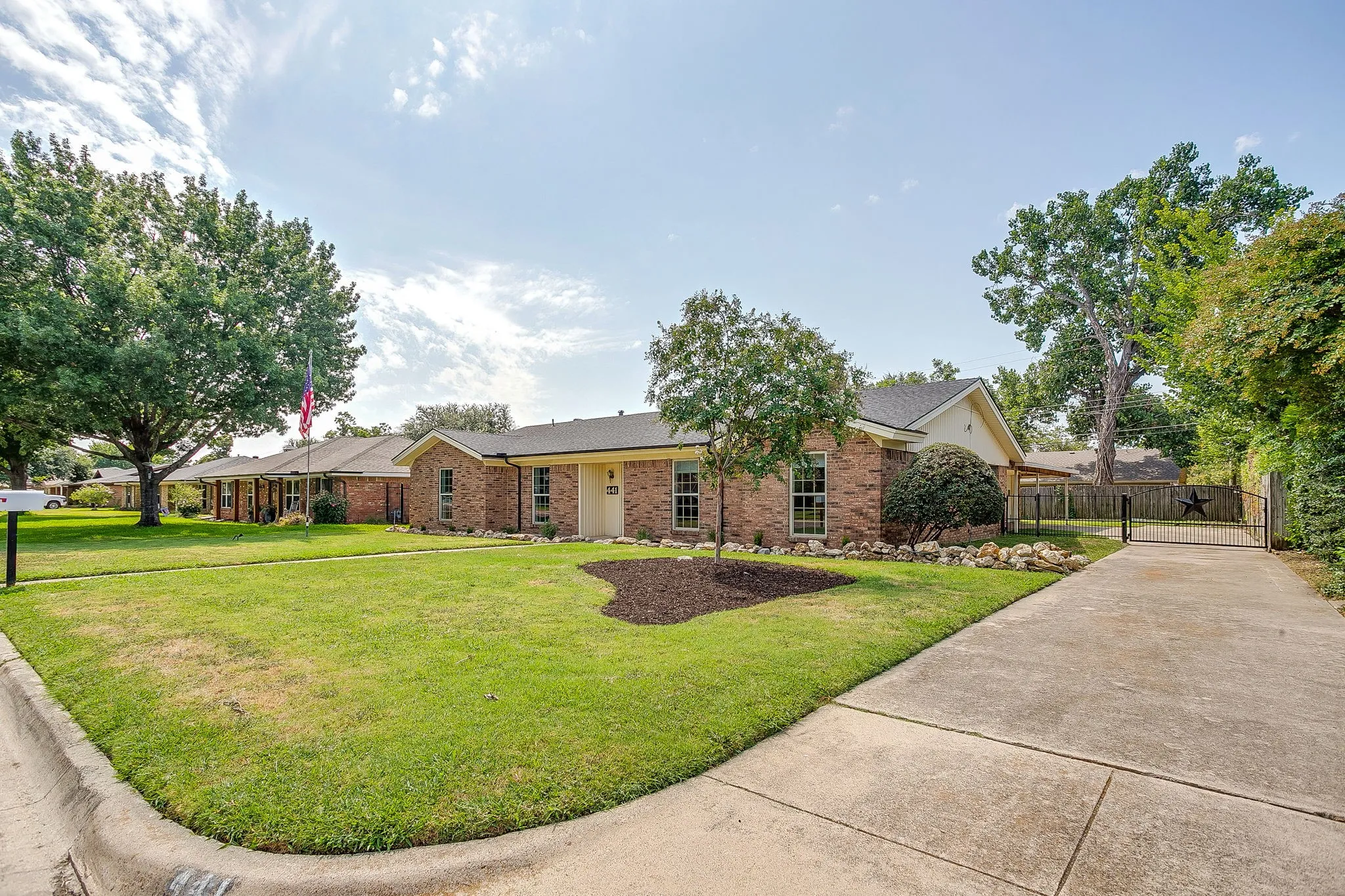 Brick exterior with a gabled roofline, mature trees, concrete driveway, and a decorative metal gate