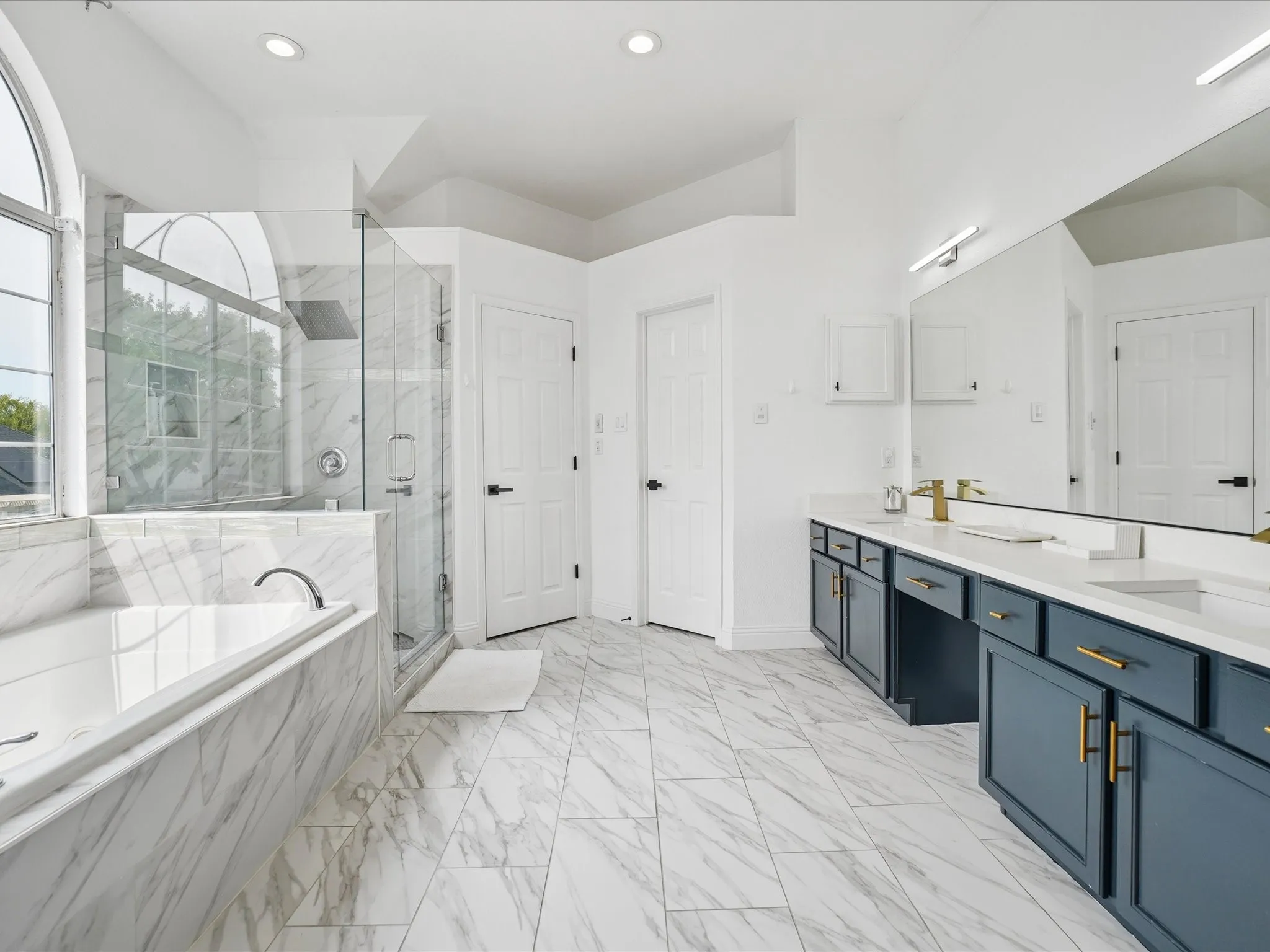 Spacious bath featuring marble-look tile flooring, an arched window, a built-in tub with marble-look surround, a glass-enclosed shower, and a dual vanity with white countertops and navy cabinetry