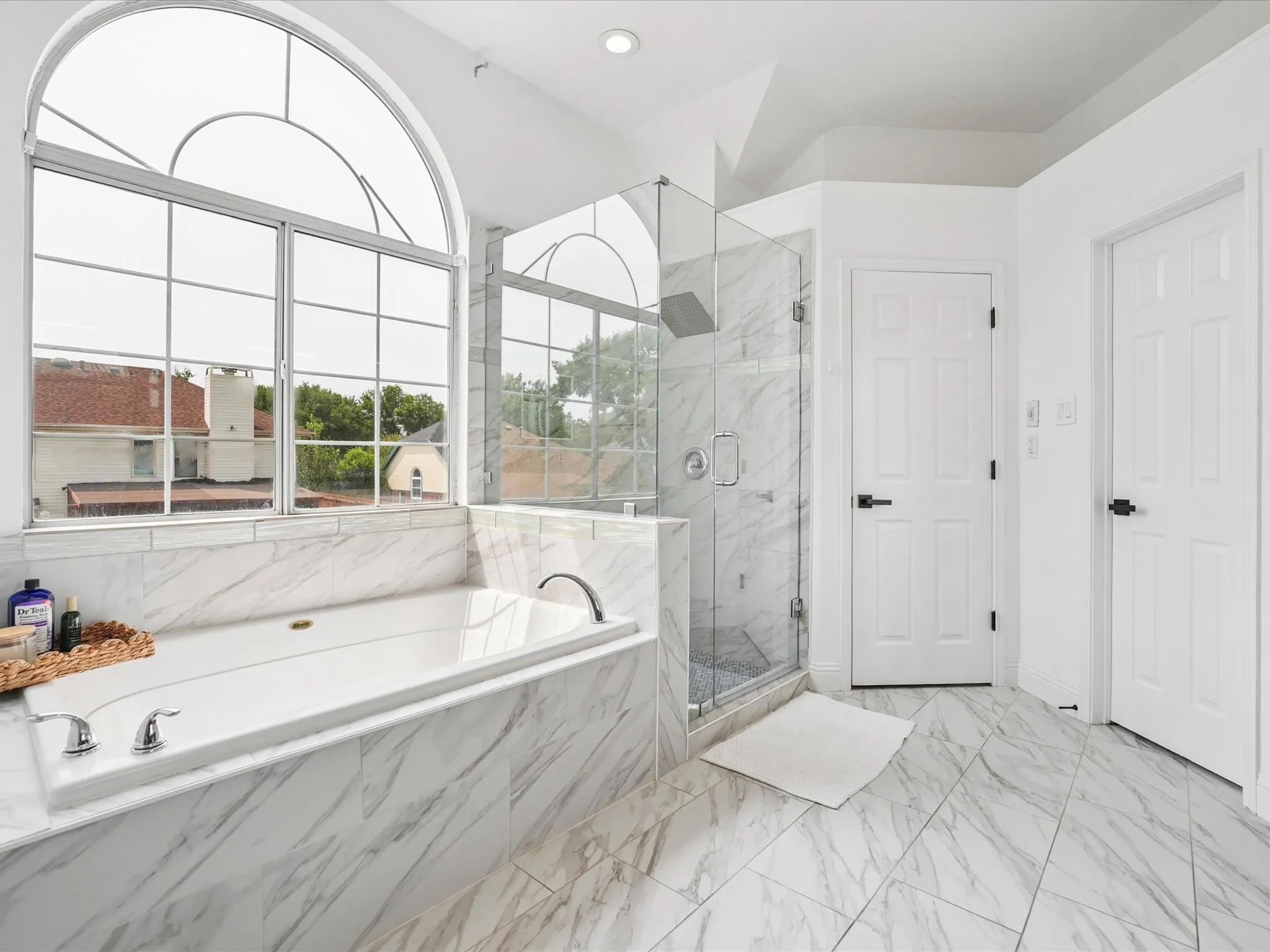 Soaking tub with chrome fixtures, surrounded by marble-style tiling, and a large arched window