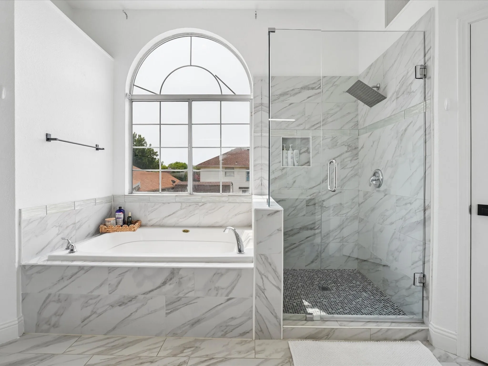 Spa-inspired master bath featuring a sculptural arched window with gridded panes, Carrara marble surround and soaking tub, and a walk-in shower with marble tile walls, frameless glass enclosure, and pebble floor