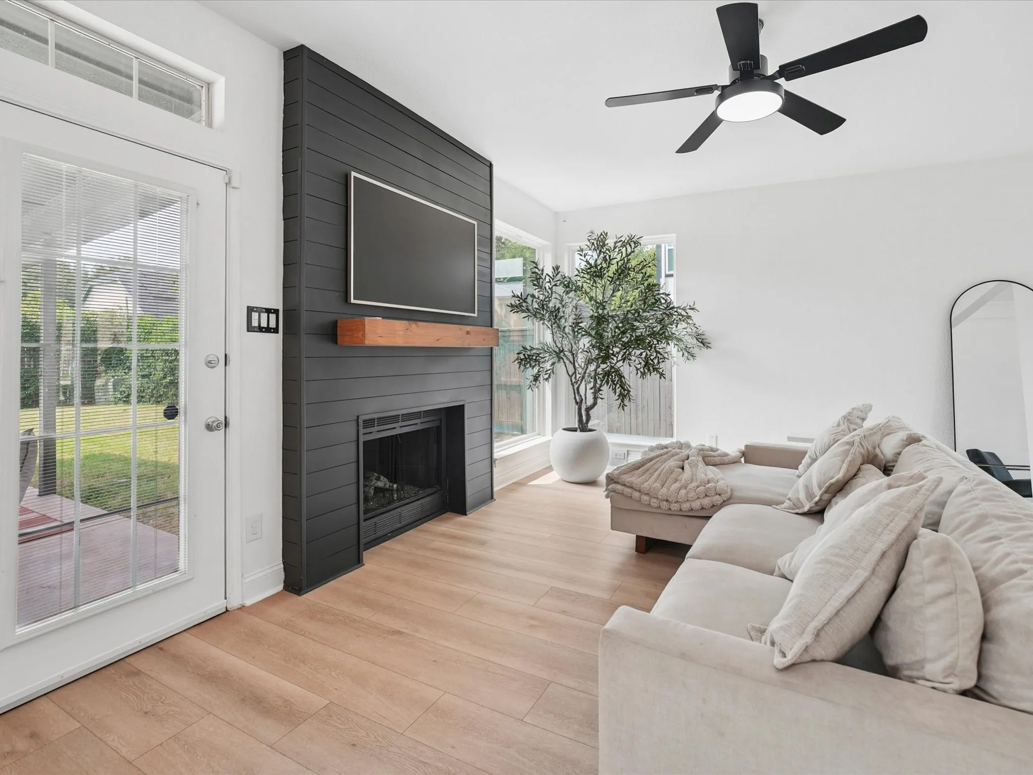 Living area featuring wood-finish flooring, a contemporary black shiplap fireplace with a wood mantel, a ceiling fan, and a glass-paned door with an upper transom window