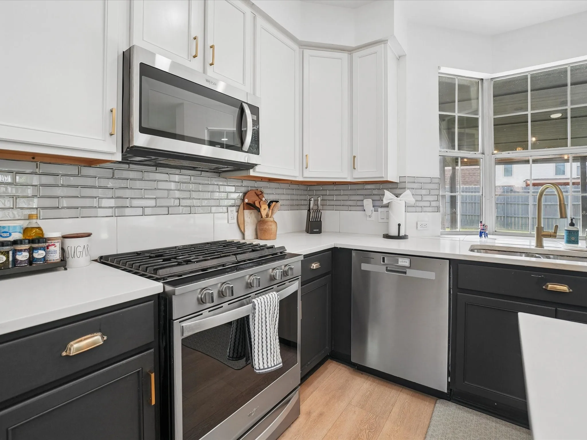 Modern kitchen featuring two-tone cabinetry with gold-finish hardware, stainless steel appliances, a white solid surface countertop, glass subway tile backsplash, and wood-finish flooring