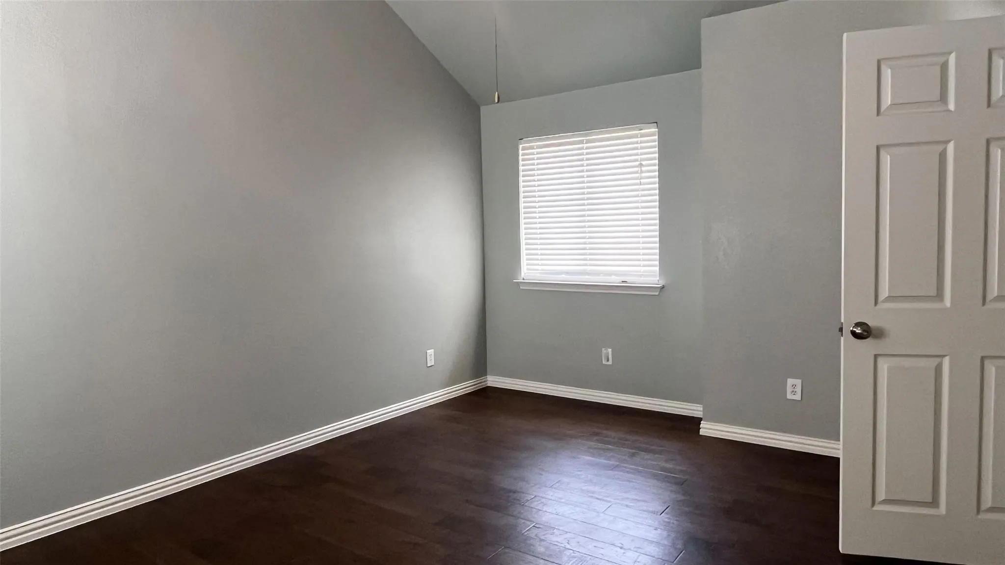Vaulted ceiling, wood-finish flooring, neutral wall tones, window with horizontal blinds, paneled door