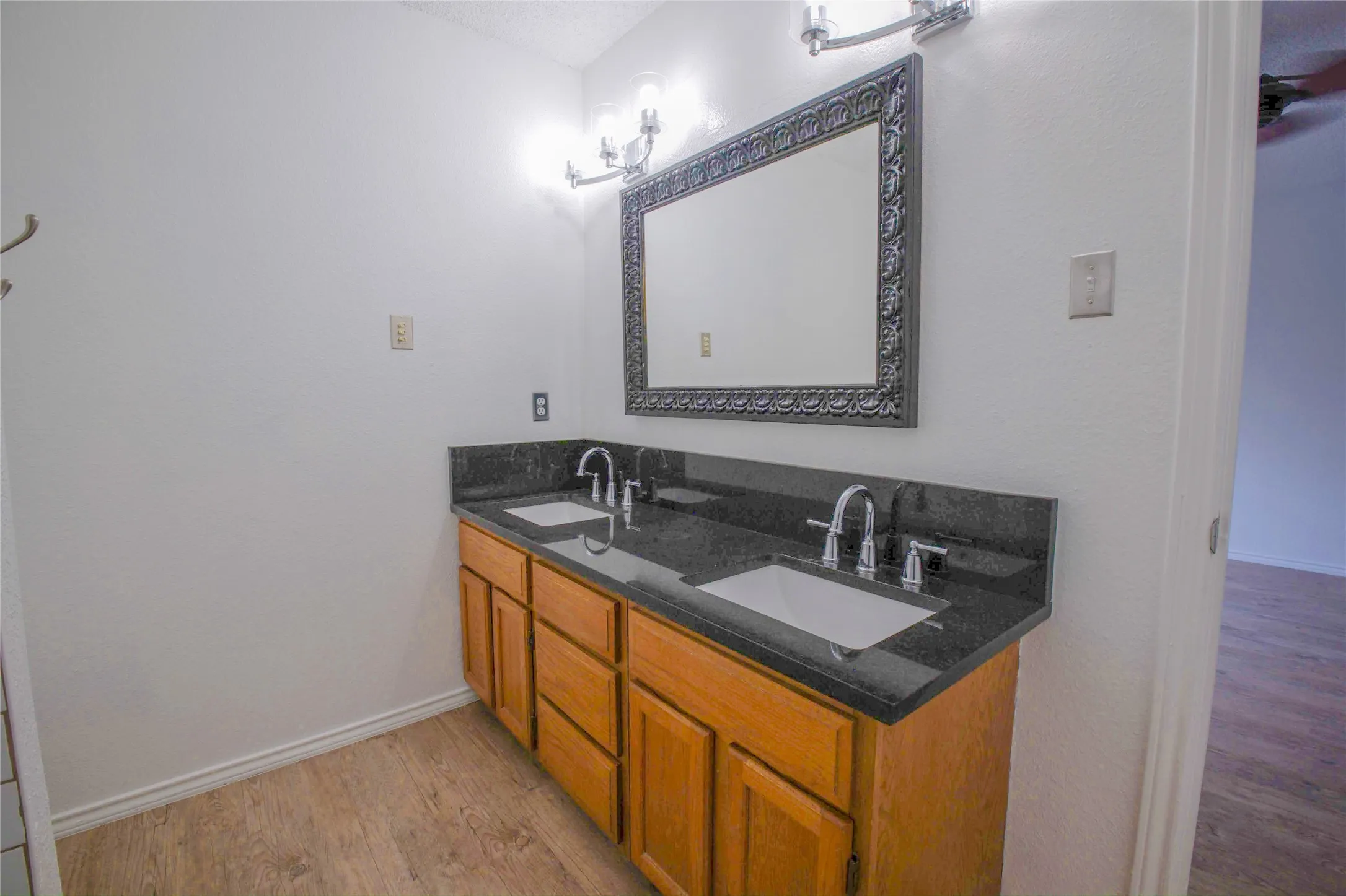 Wood-finish flooring with a dual vanity featuring a dark countertop, undermount sinks, and chrome faucets
