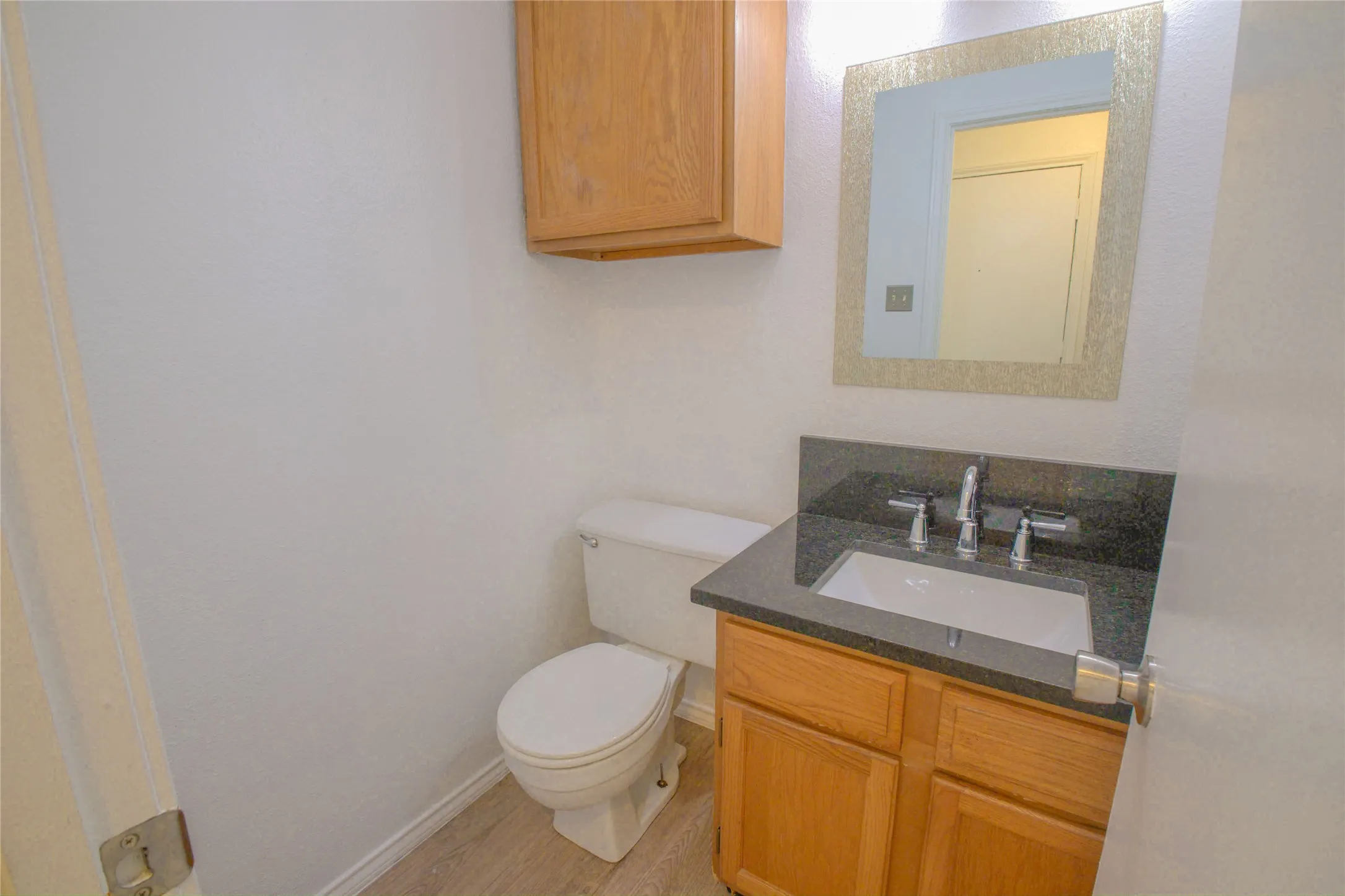 Wood-finish flooring, a toilet, a vanity with a dark countertop and integrated sink, a mirrored medicine cabinet, and a wall-mounted light fixture