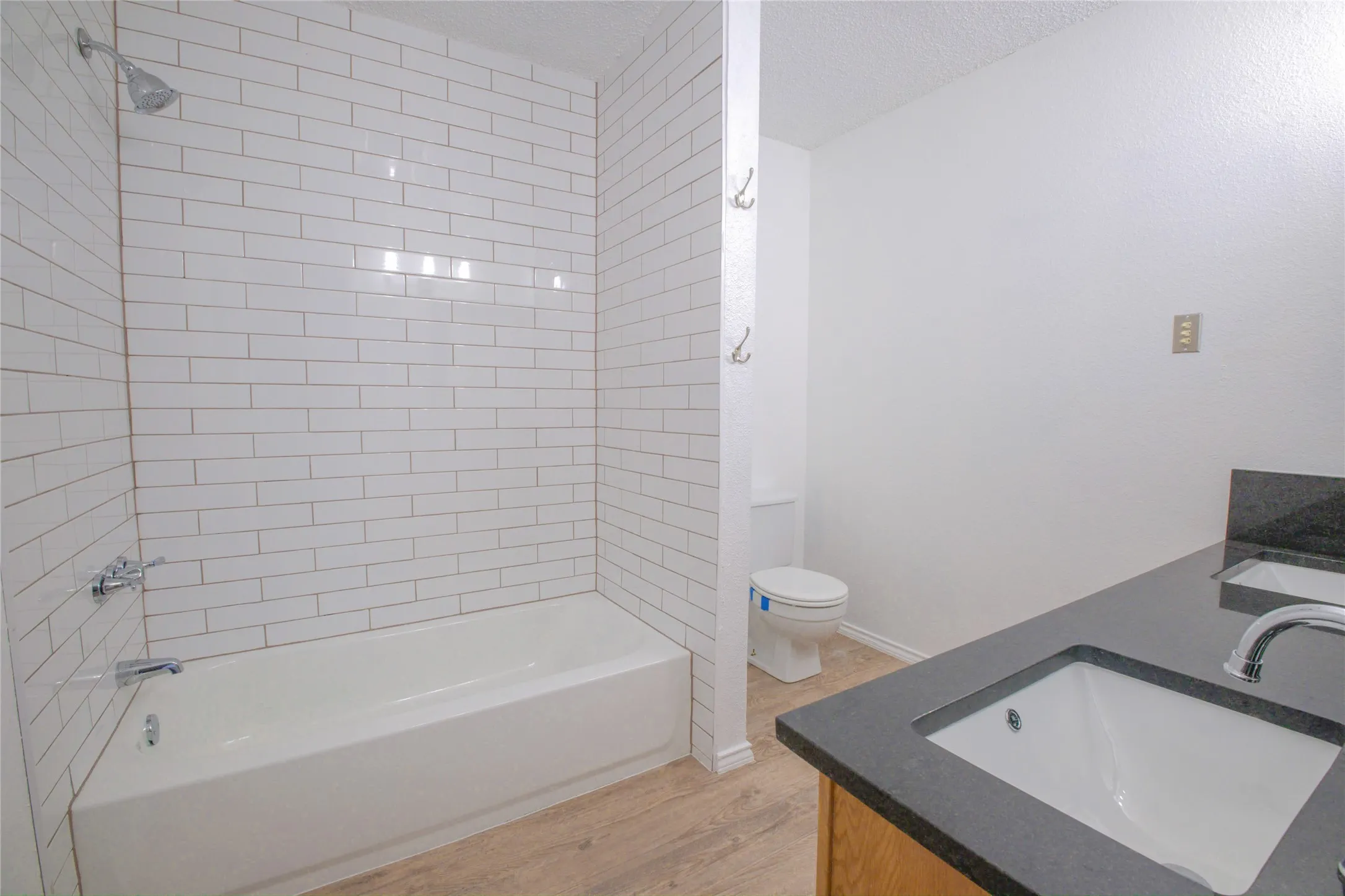 Bathtub with subway tile surround, chrome shower fixtures, wood-finish flooring, and a dark countertop vanity with an integrated sink