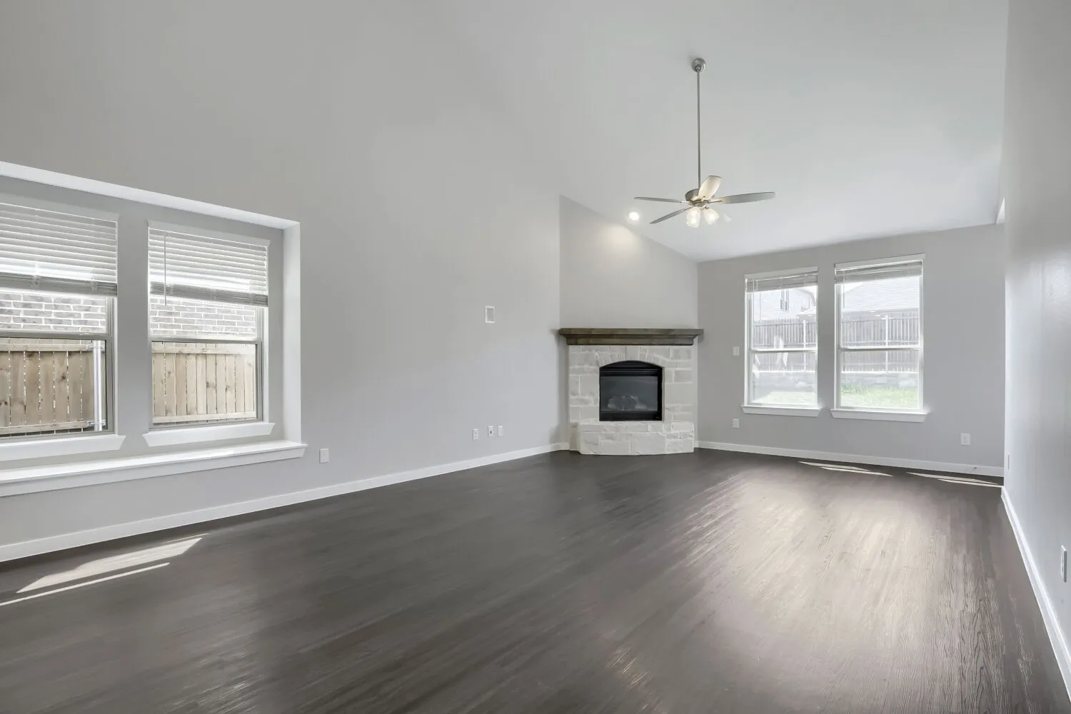 Vaulted ceiling, stone-surround fireplace with wood mantel, dark wood-finish flooring, ceiling fan, and multiple windows