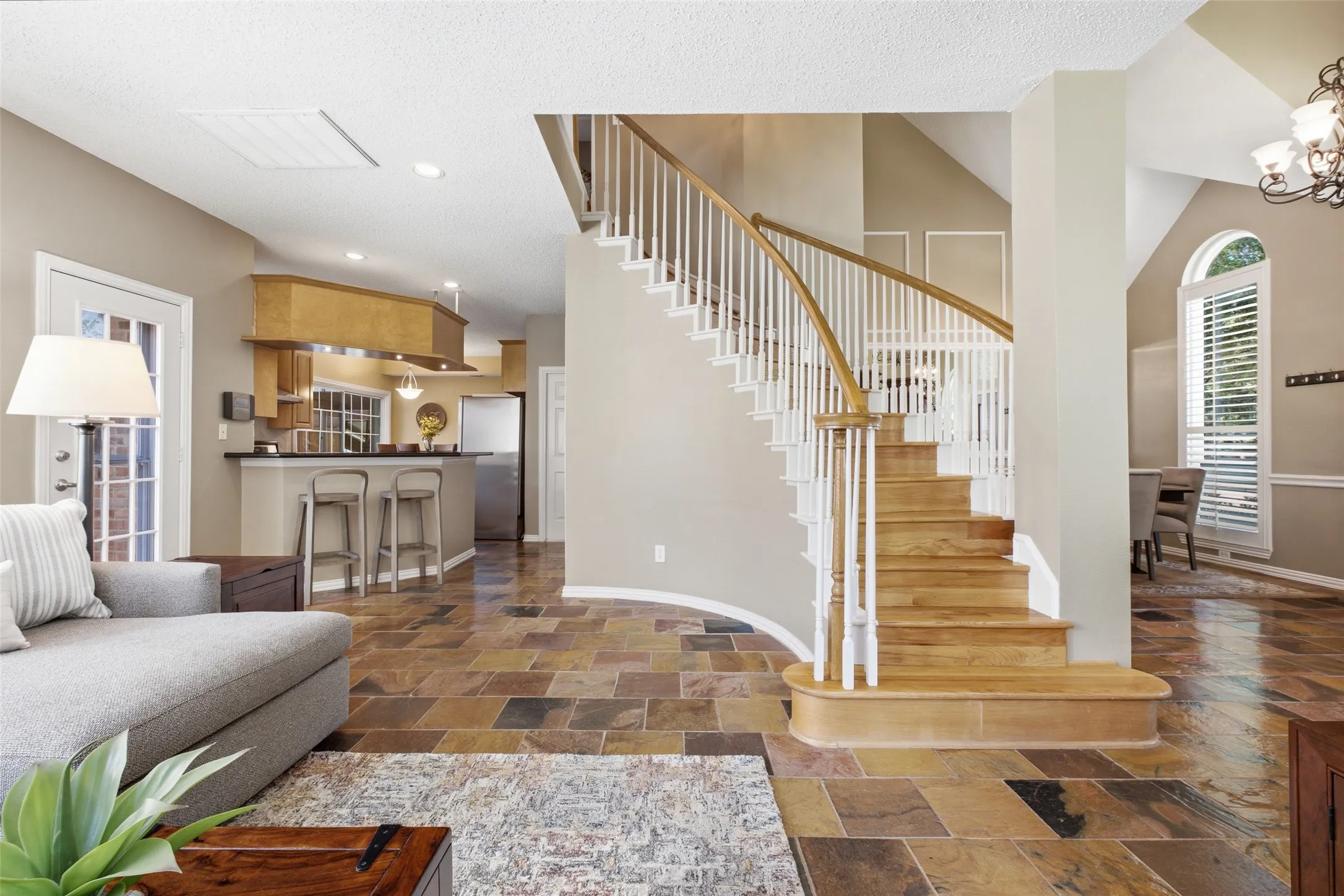 Grand open floor plan with a sweeping curved staircase, slate tile floors, and sightlines connecting the family room, kitchen, and formal dining — space and elegance in perfect balance.