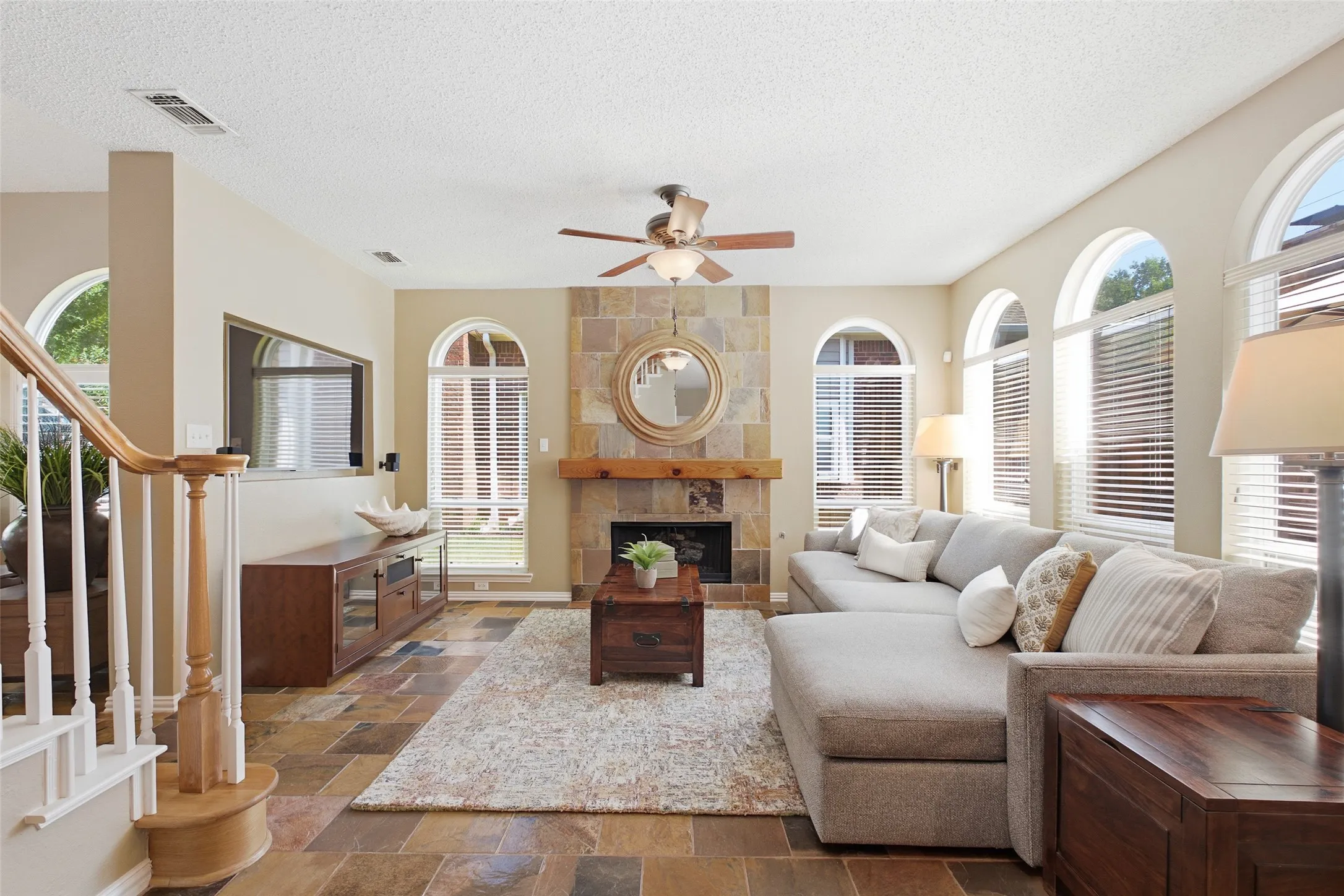 Spacious family room with a floor-to-ceiling slate tile fireplace, wood beam mantle, oversized sectional, and abundant natural light flooding in through triple arched windows.
