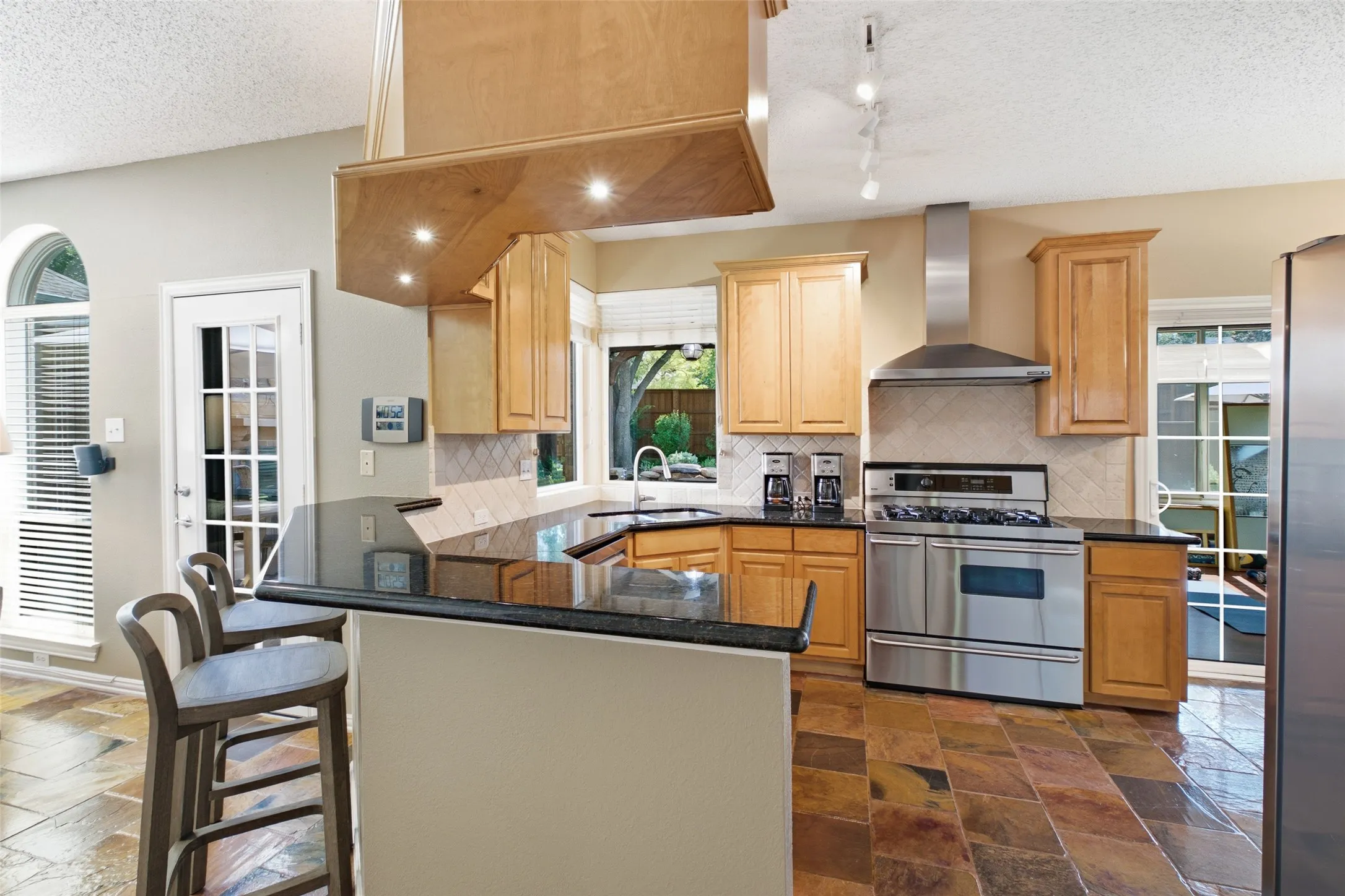 Kitchen featuring maple cabinetry, black granite countertops, a custom wood-wrapped range hood, stainless steel appliances, gas range, and breakfast bar seating.