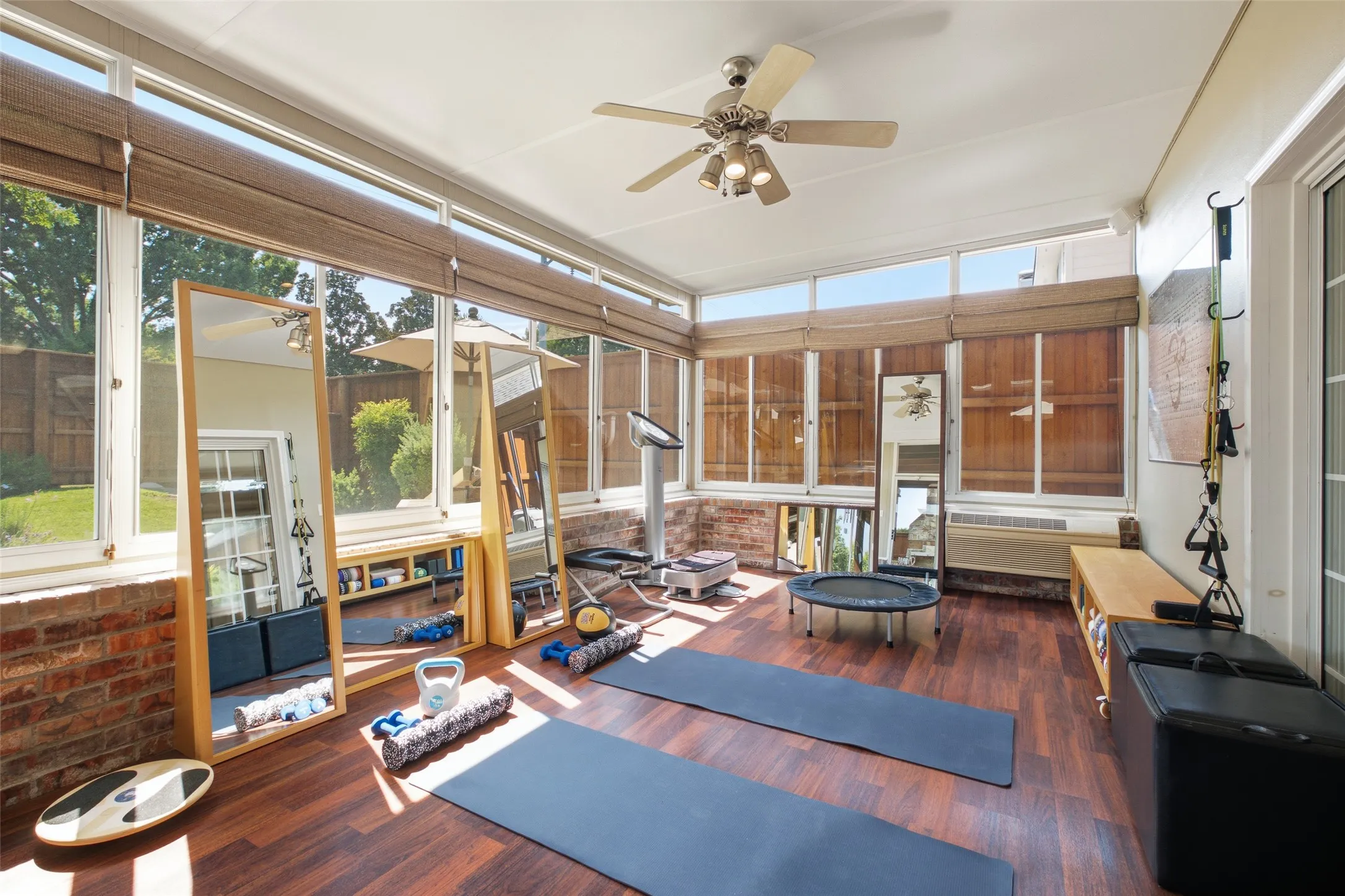 Versatile sunroom currently used as a private home gym, featuring hardwood floors, walls of windows, abundant natural light, and direct access to the backyard.