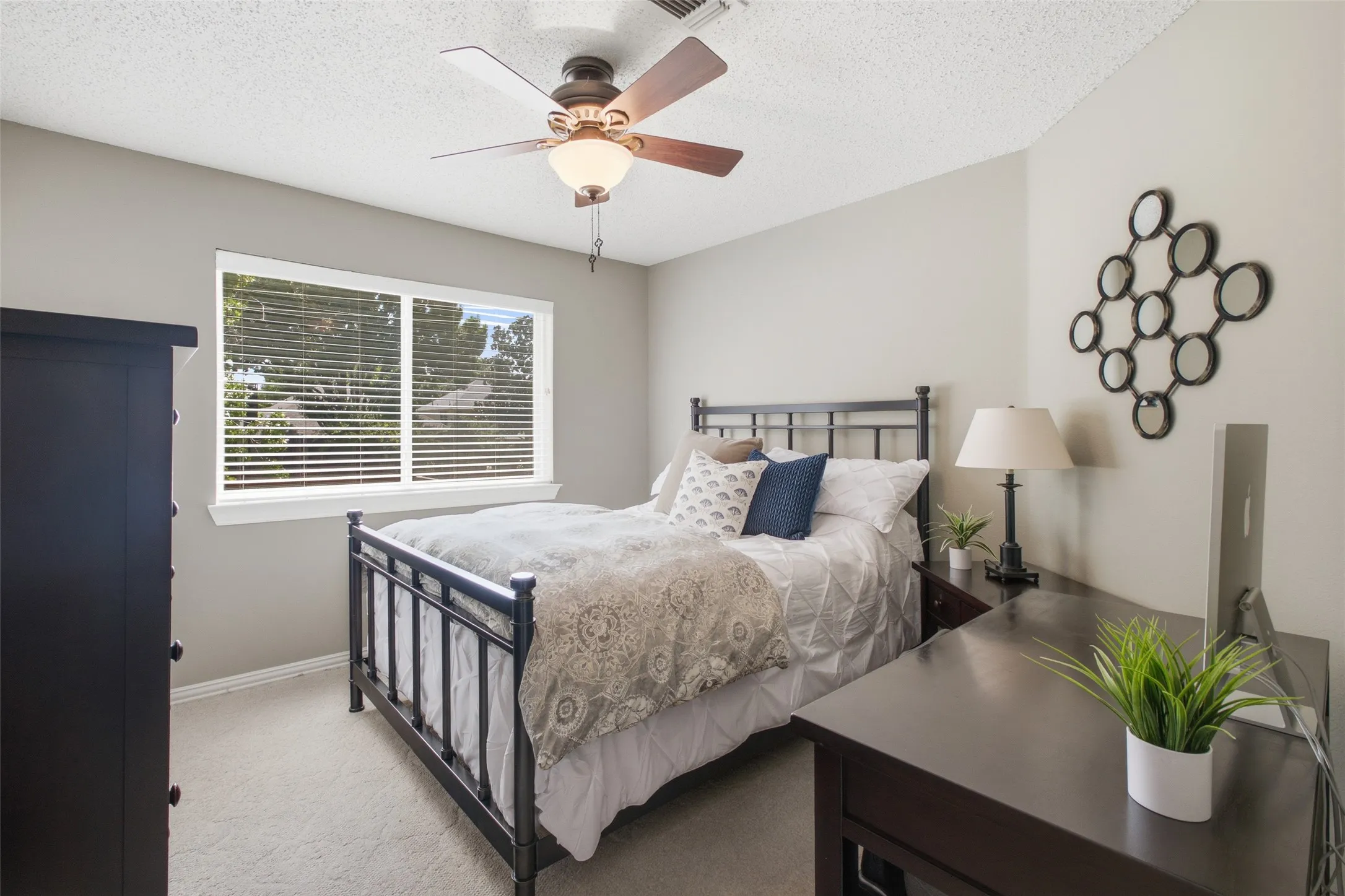 Secondary bedroom with an iron bed frame, ceiling fan, carpet, and a window overlooking mature trees.