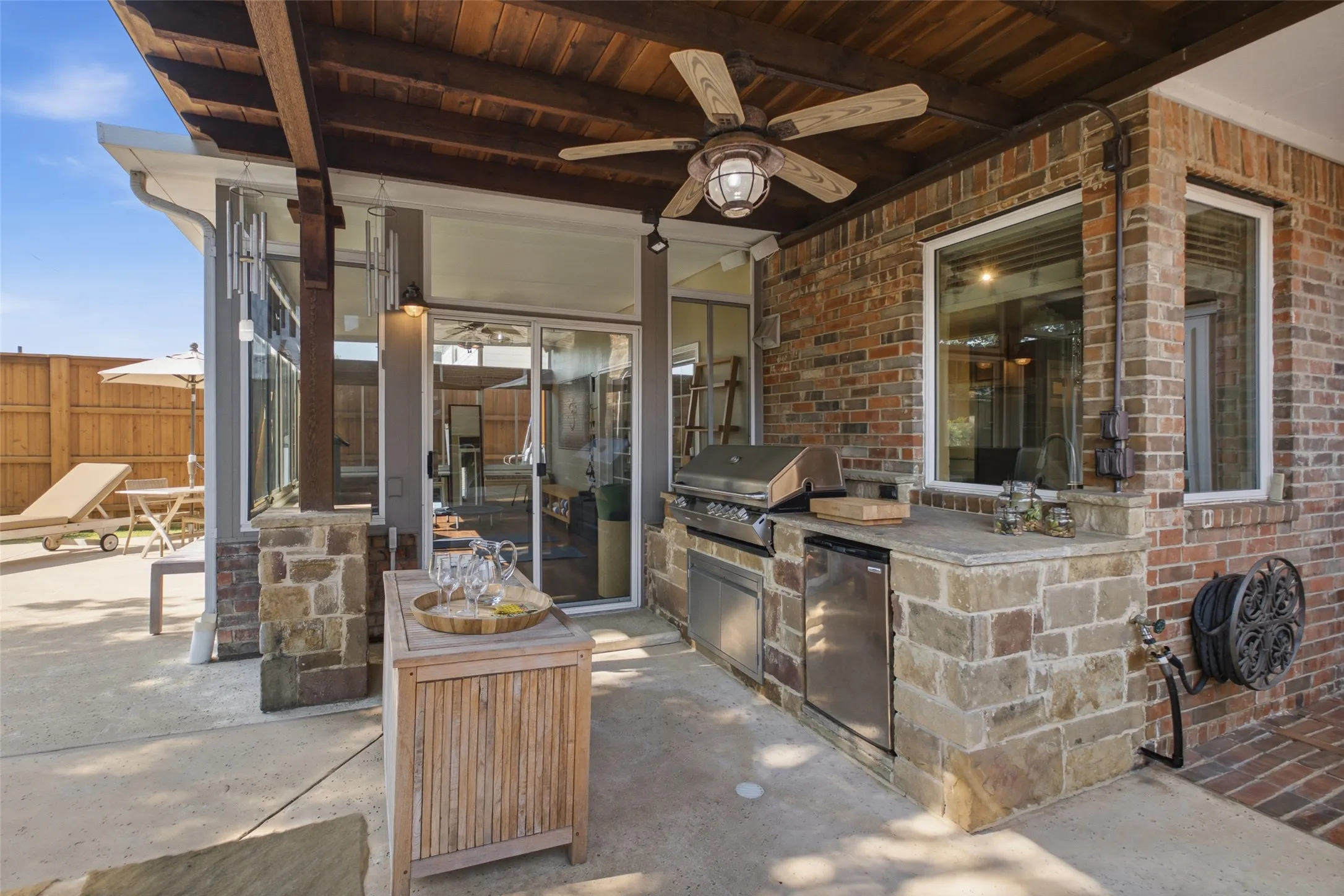 Covered outdoor kitchen with built-in gas grill, mini refrigerator, stone countertops, cedar ceiling with fan, and direct views into the sunroom — outdoor entertaining at its finest.