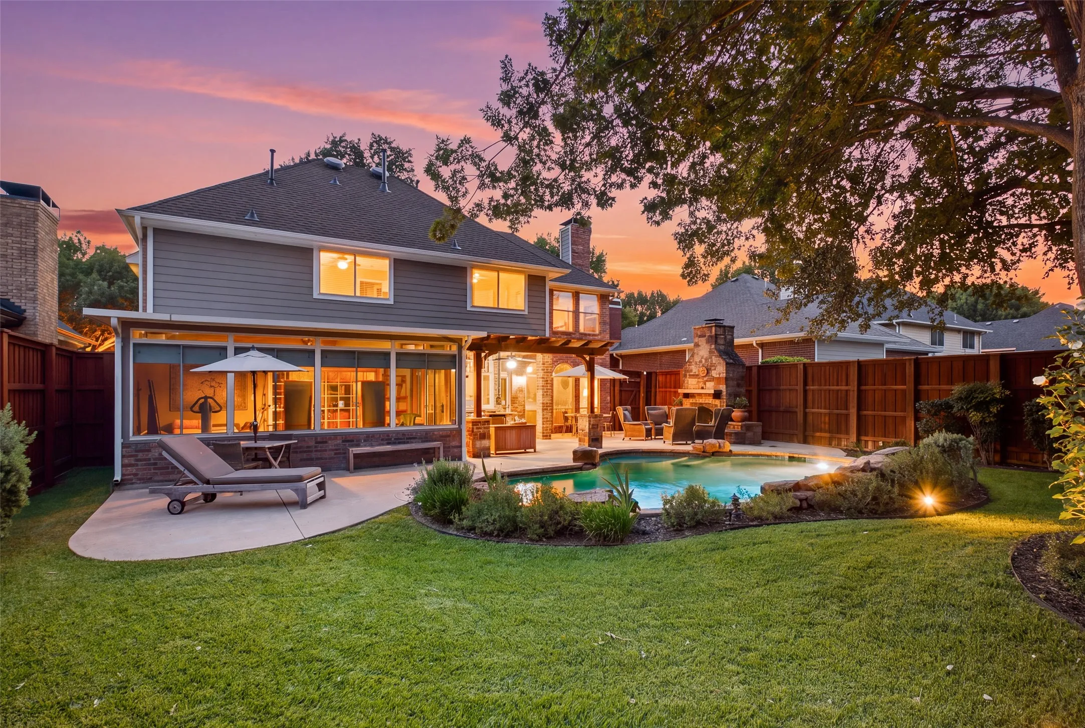 Sweeping twilight view of the backyard showcasing the sunroom, covered patio, sparkling pool, outdoor fireplace, and beautifully manicured lawn — it all comes together here.