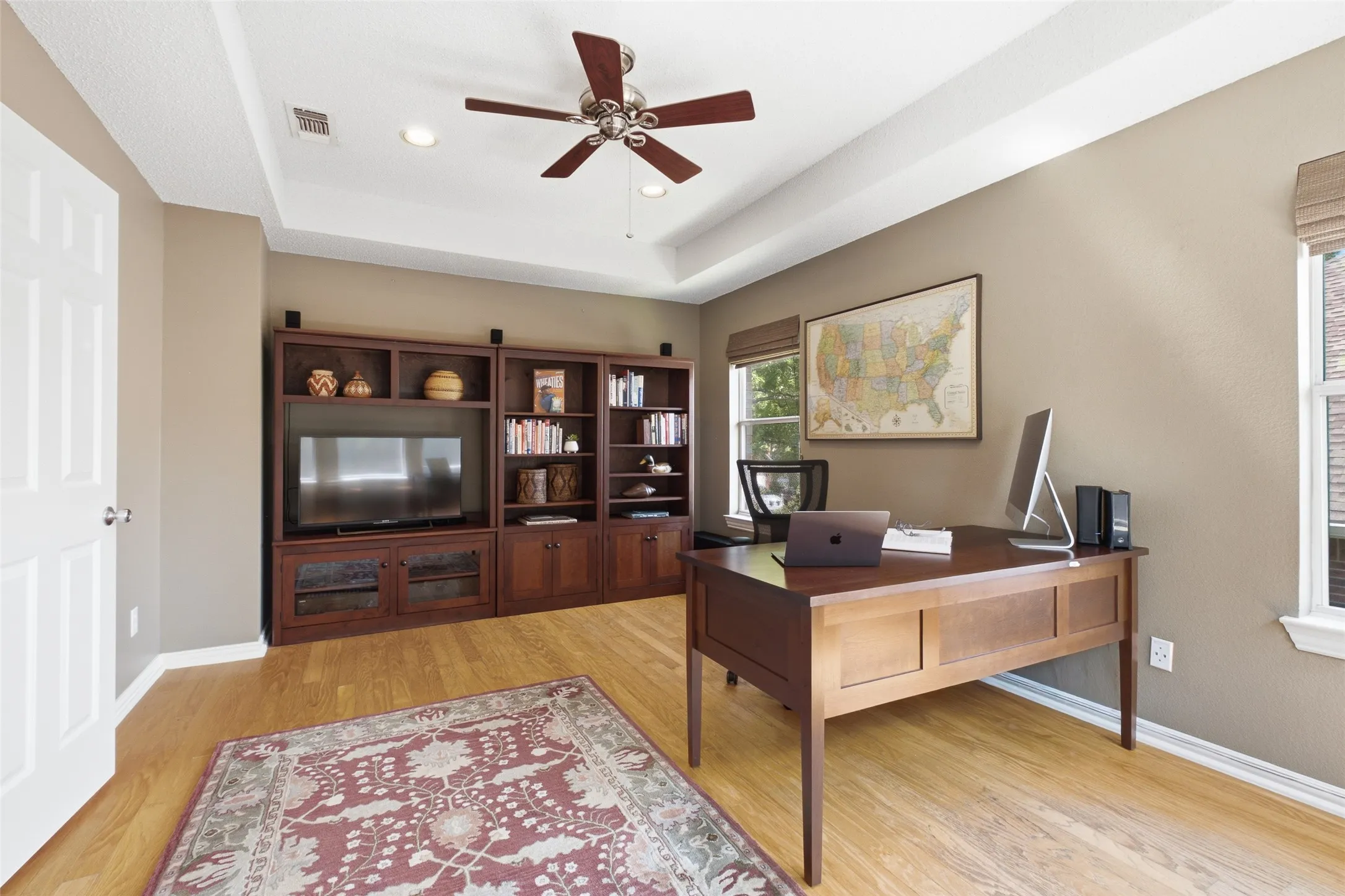 Spacious home office with built-in bookcase, wall-mounted TV, executive desk, tray ceiling, and hardwood floors — a distinguished and functional workspace.