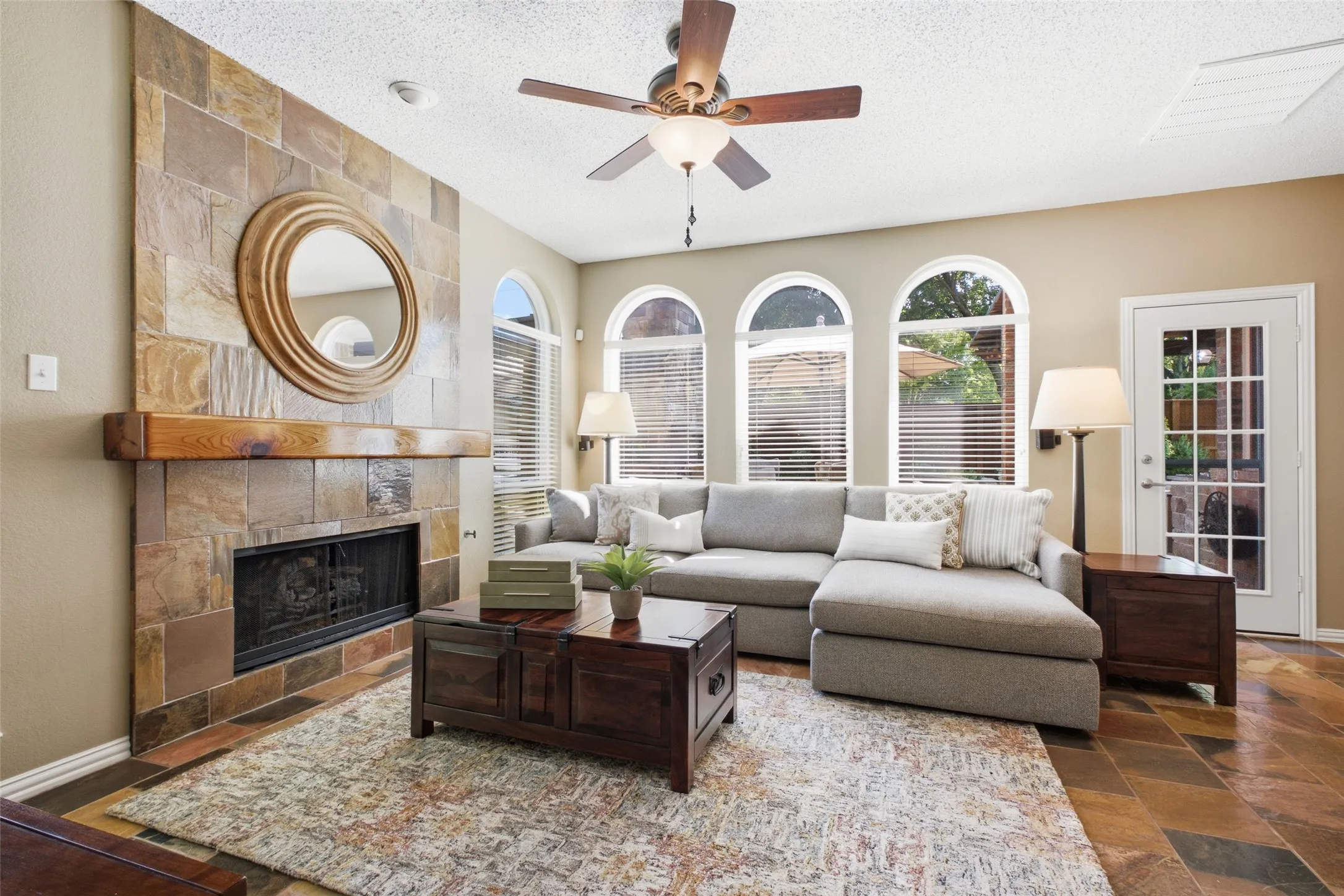 Family room showcasing the striking slate stone fireplace with wood mantle, triple arched windows overlooking the backyard, and access to the patio.
