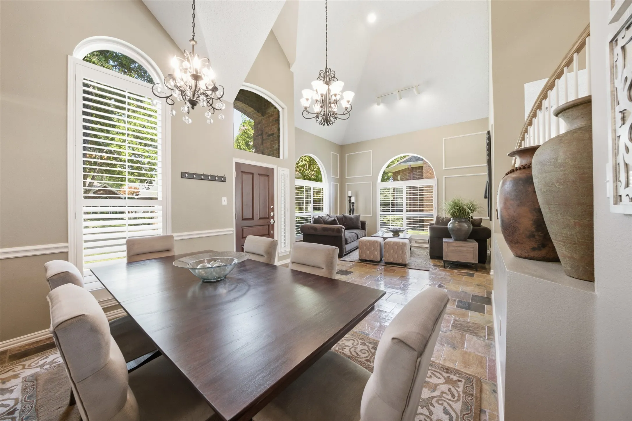 Soaring vaulted formal dining room with dual chandeliers, a large dark wood dining table, arched windows with shutters, and open sightlines to the formal living room.