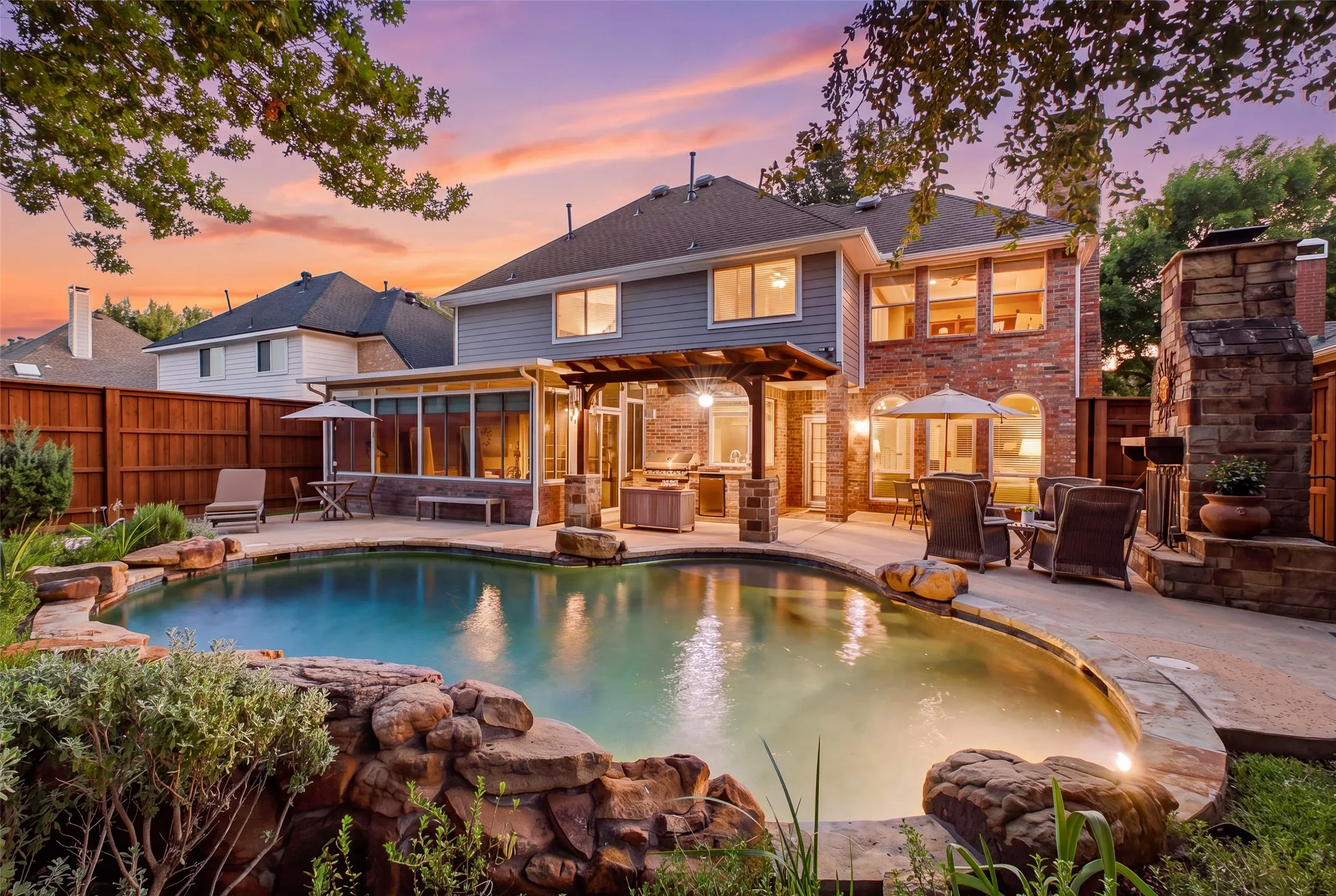 Stunning resort-style backyard at twilight — freeform pool with natural rock waterfall, covered pergola, outdoor kitchen, and stone fireplace create the ultimate private retreat.