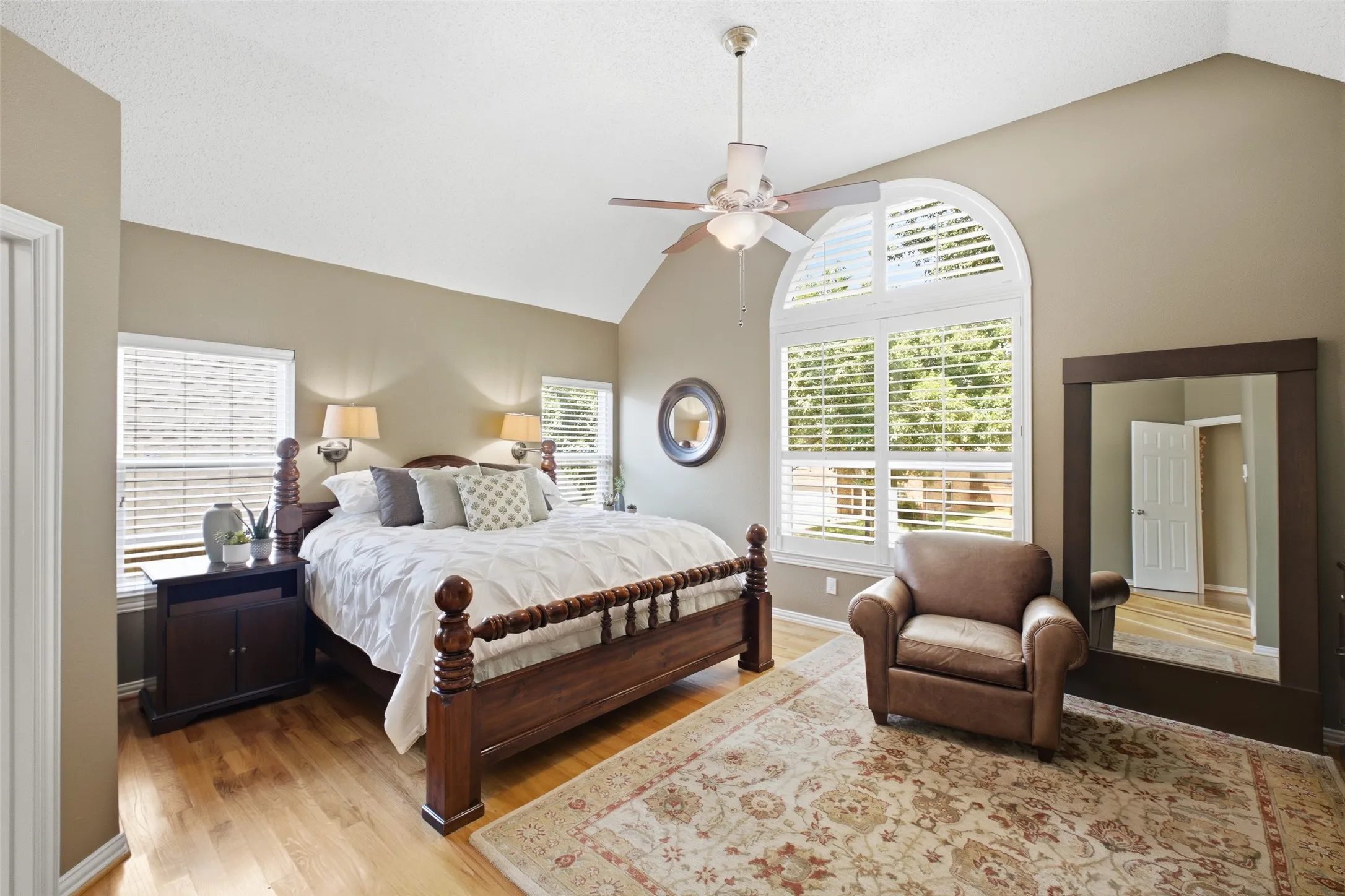 Luxurious primary suite with vaulted ceilings, a dramatic arched window, four-poster bed, plantation shutters, and gleaming hardwood floors.
