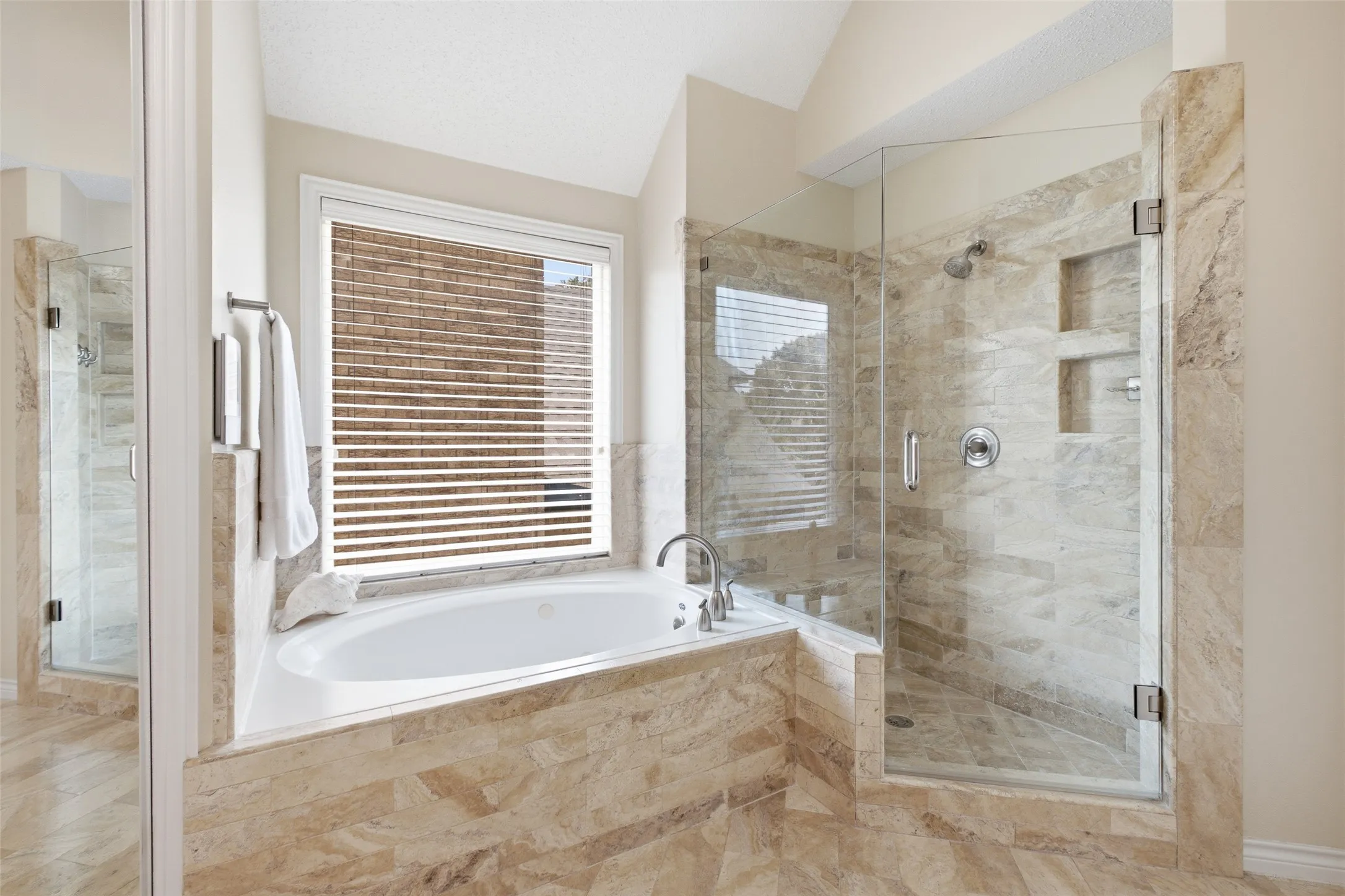 Spa-inspired primary bathroom featuring a soaking tub, frameless glass walk-in shower, and travertine tile surround — a true daily retreat.