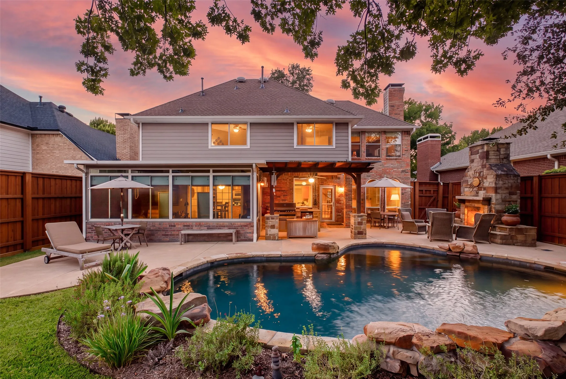 Breathtaking backyard at sunset featuring a sparkling pool with rock waterfall, cedar-ceiling covered living area, outdoor fireplace, and lush landscaping designed for effortless entertaining.