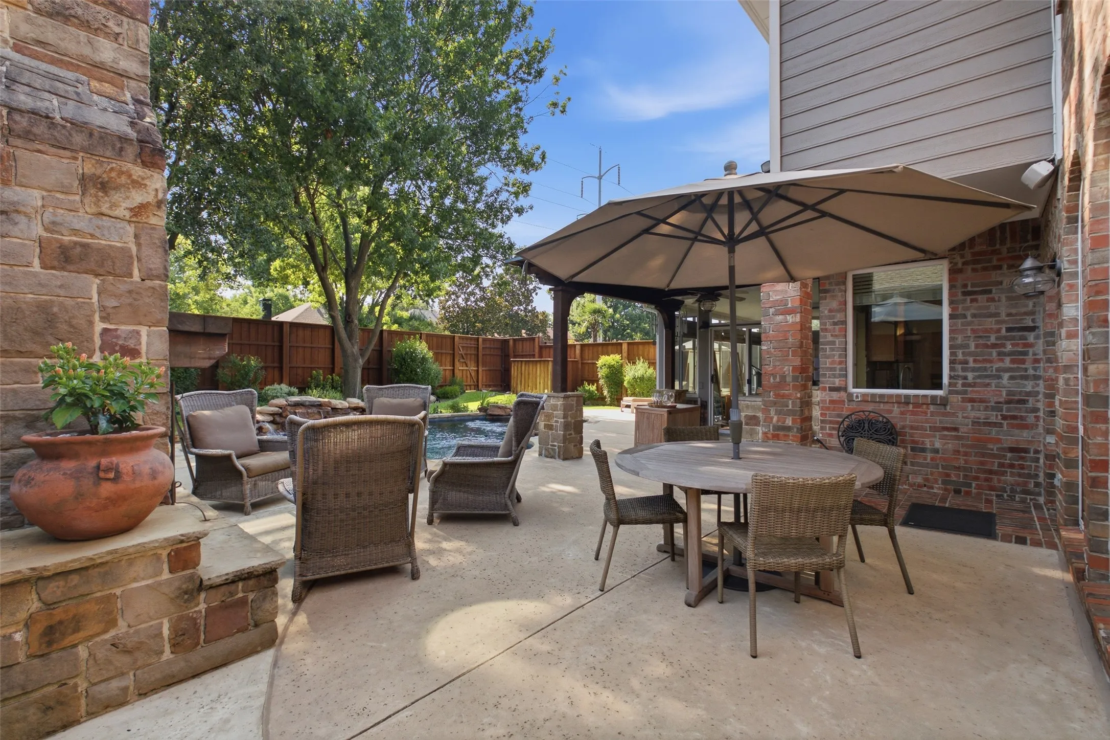 Expansive patio with multiple outdoor living zones — wicker lounge seating, alfresco dining under a market umbrella, and pool views surrounded by lush landscaping.