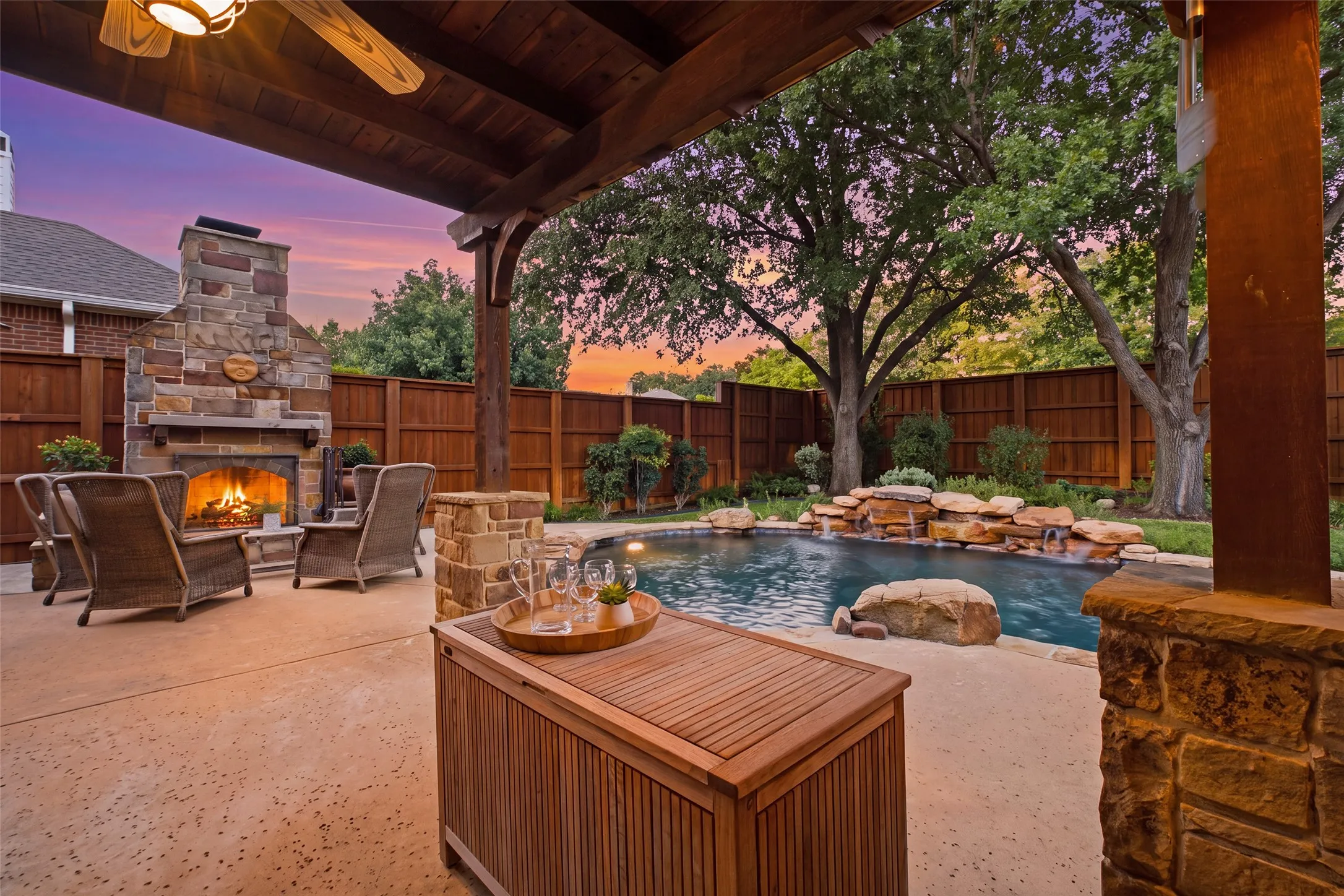 Magical backyard at sunset — the covered pergola with a crackling stone fireplace, sparkling pool with rock waterfall, and towering mature trees create an unmatched outdoor escape.