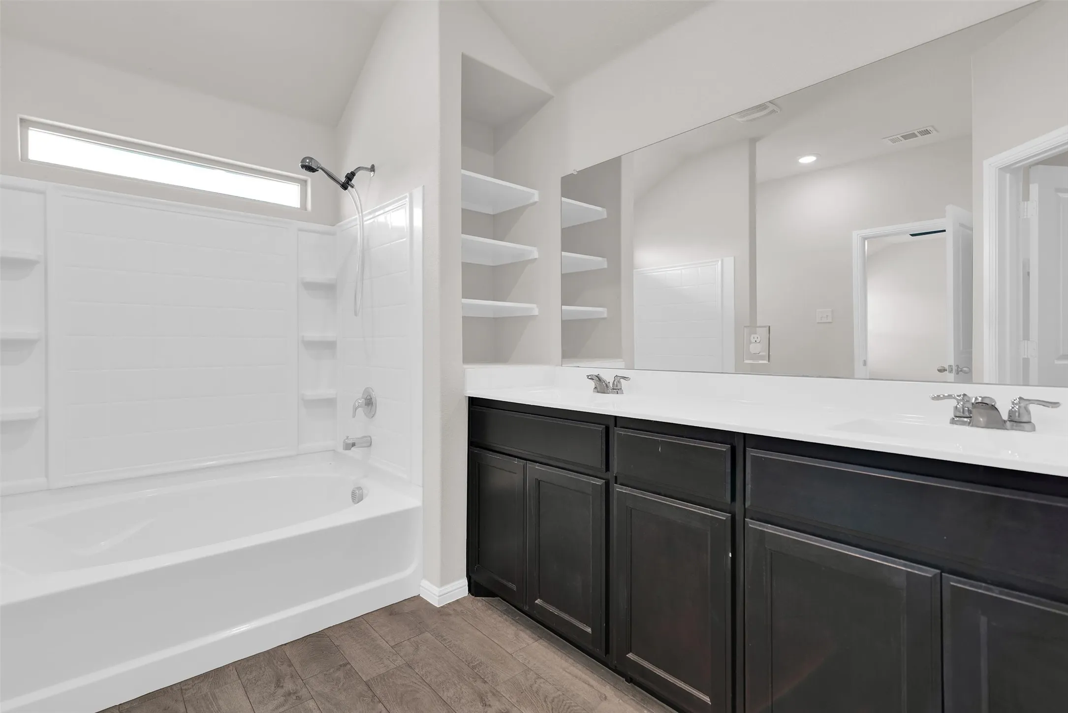 Bathroom featuring a white tub and shower combination, dark wood-finish vanity with dual sinks, a large mirror, and open shelving