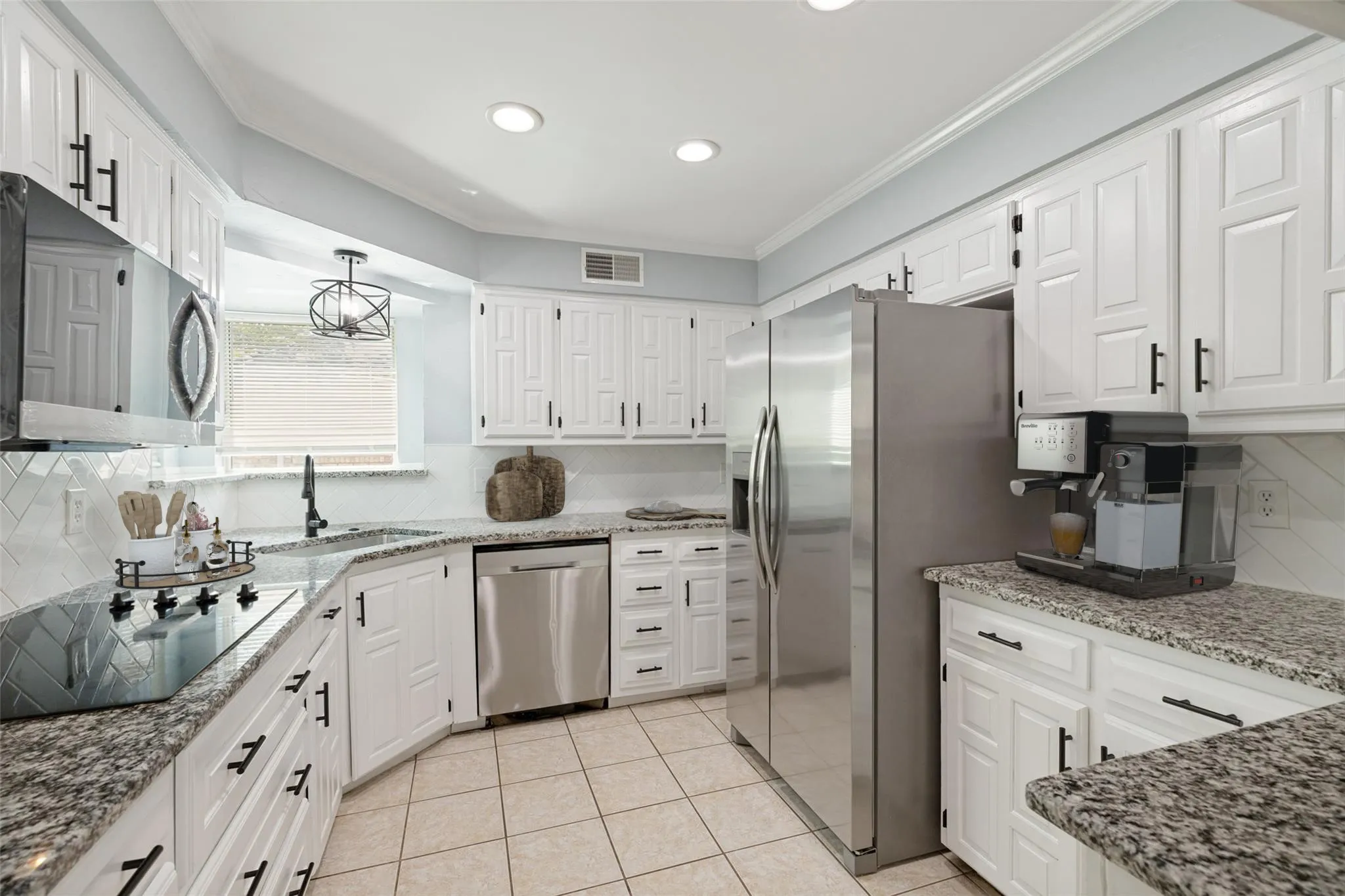 This bright and well-organized kitchen showcases a beautifully coordinated design with white cabinetry, black hardware, and a new built-in microwave that together create a crisp, modern look. Granite countertops pair seamlessly with the diagonal tile backsplash, adding texture and visual interest across the entire workspace. Stainless steel appliances, including the refrigerator, dishwasher, microwave, and cooktop, provide a polished, contemporary finish. A window above the sink brings in natural light and offers a pleasant view outside, while recessed lighting and a modern ceiling fixture ensure the space stays well-lit and inviting. The neutral tile flooring ties the room together with a warm, cohesive feel that complements the kitchen’s clean, updated presentation.