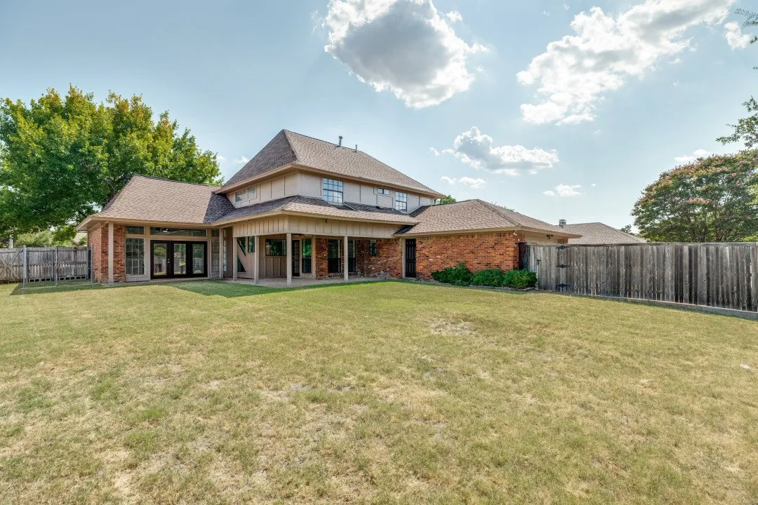 This photo showcases the home’s expansive backyard, made possible by its large lot size, offering a wide stretch of green space perfect for outdoor living, play, or future personalization. The covered patio with a ceiling fan provides a comfortable shaded area for relaxing or entertaining, seamlessly connecting indoor and outdoor spaces. The home’s attractive brick-and-siding exterior, large windows, and glass doors create a bright, inviting backdrop while enhancing appeal from every angle. Fully fenced and bordered by mature greenery, this backyard delivers both privacy and plenty of room to enjoy.