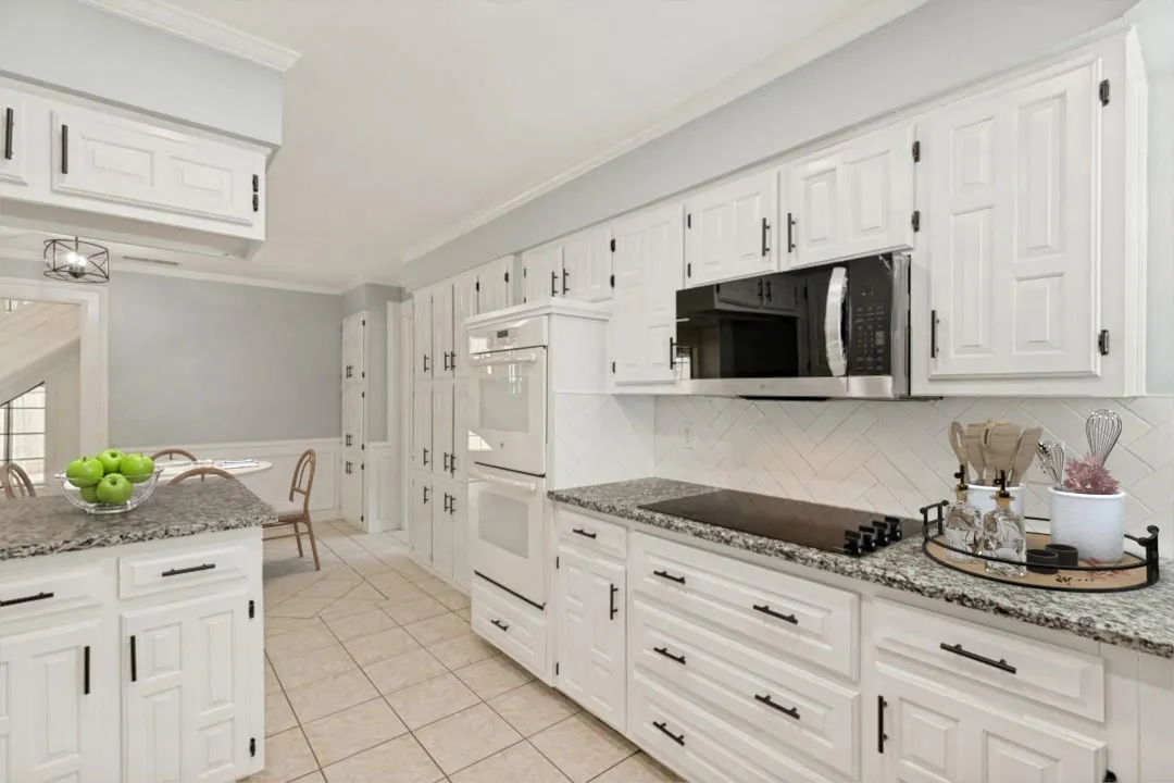 This elegant kitchen angle highlights the herringbone tile backsplash, adding a refined, textured focal point behind the cooktop. The gray granite countertops pair beautifully with the crisp white cabinetry and black hardware, creating a modern, cohesive look. The built-in oven, black glass cooktop, and integrated microwave offer a streamlined appliance layout that enhances both function and style. Light tile flooring keeps the space bright, while the soft wall color and white trim add a clean, contemporary finish. A glimpse of the staircase beyond the kitchen provides a sense of connection to the rest of the home and showcases the thoughtful flow of the layout.